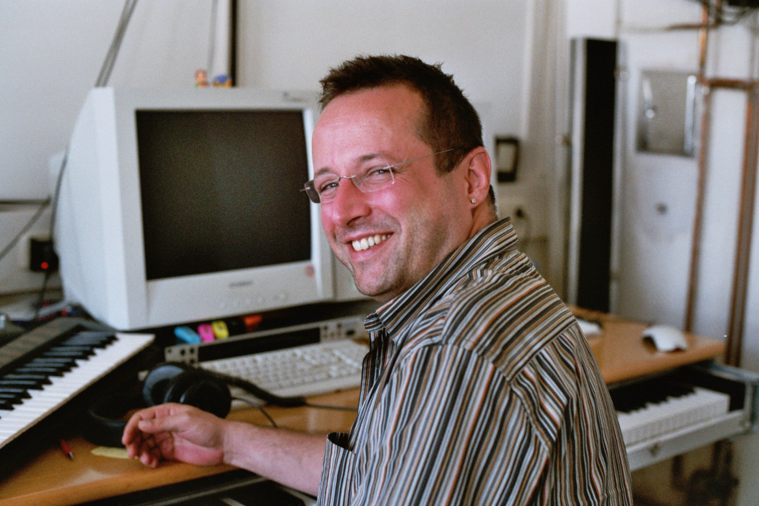 Man smiling while working in a home music studio with keyboards, headphones, and computermonitor.
