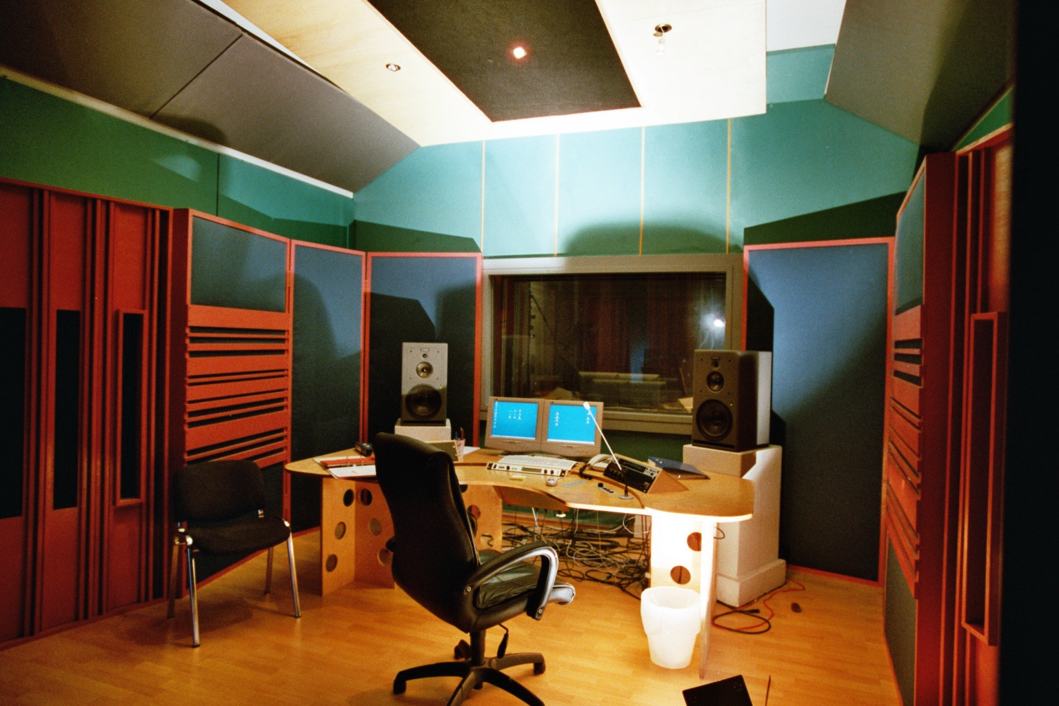 Professional recording studio with acoustically treated walls, soundproof window, control desk, dual computer monitors, audio speakers, and ergonomic chair.