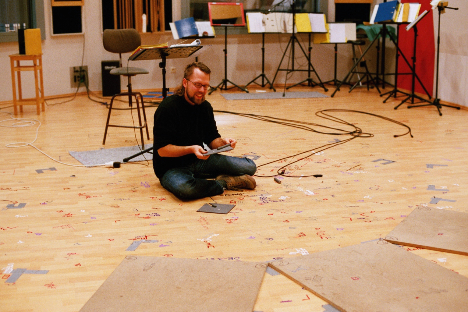 Man smiling on the floor, holding a tablet, surrounded by music stands, cables, and written notes on the wooden floor, in a recording studio.