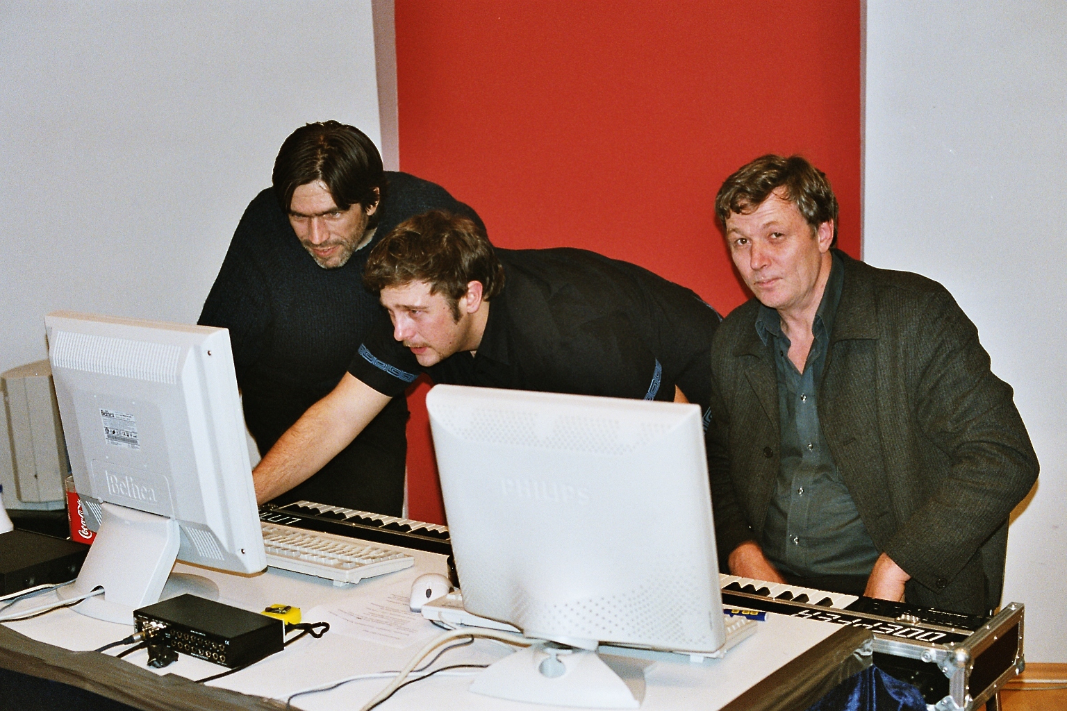 Three men collaborating on music production, leaning over computers and keyboards in a studio with a red and white wall.