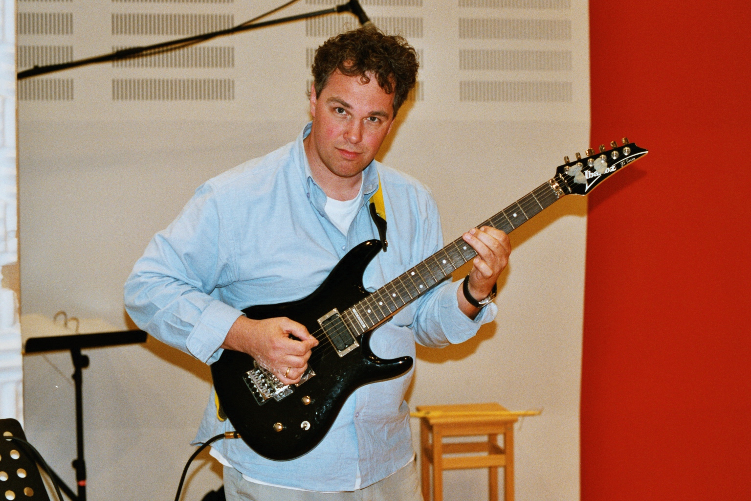 Man in blue shirt playing a black Ibanez electric guitar in a music studio.