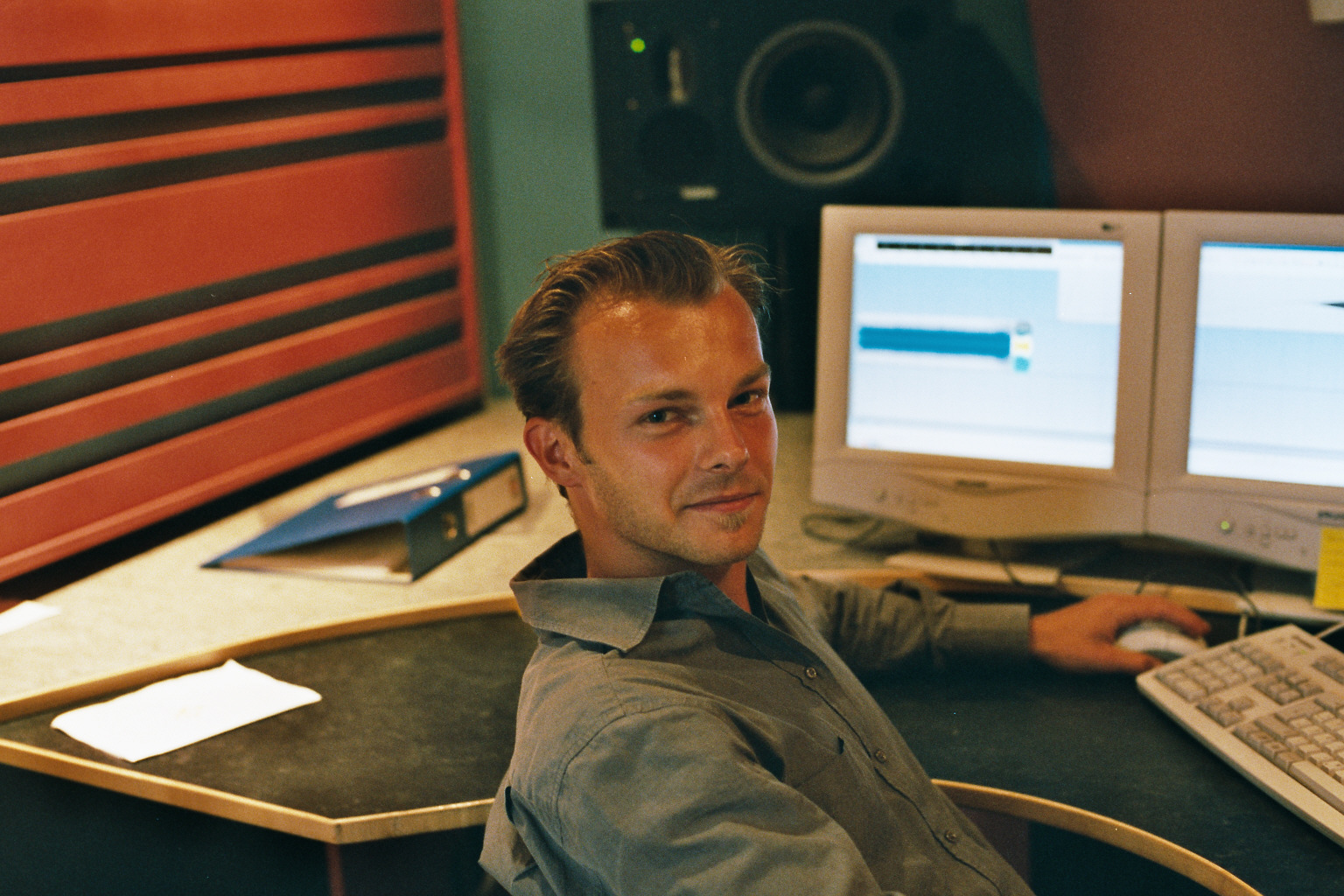 Young man in a gray shirt working on two computer monitors in a recording studio, with sound panels and audio equipment in the background.