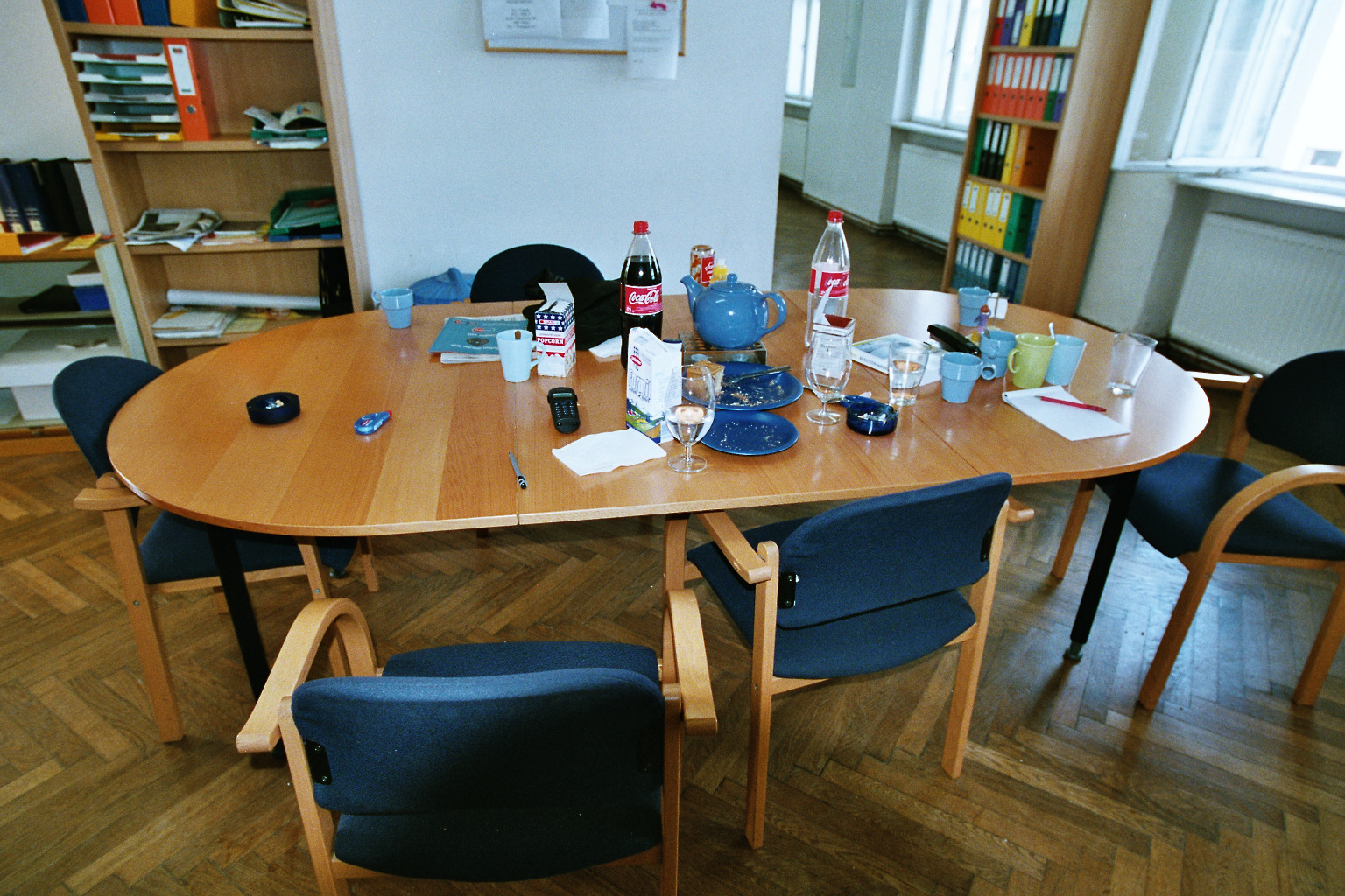 Conference table with messy items including Coca-Cola bottles, a blue teapot, empty plates, cups, glasses, a popcorn box, and a landline phone, surrounded by several blue office chairs in a room with wood flooring and bookshelves.