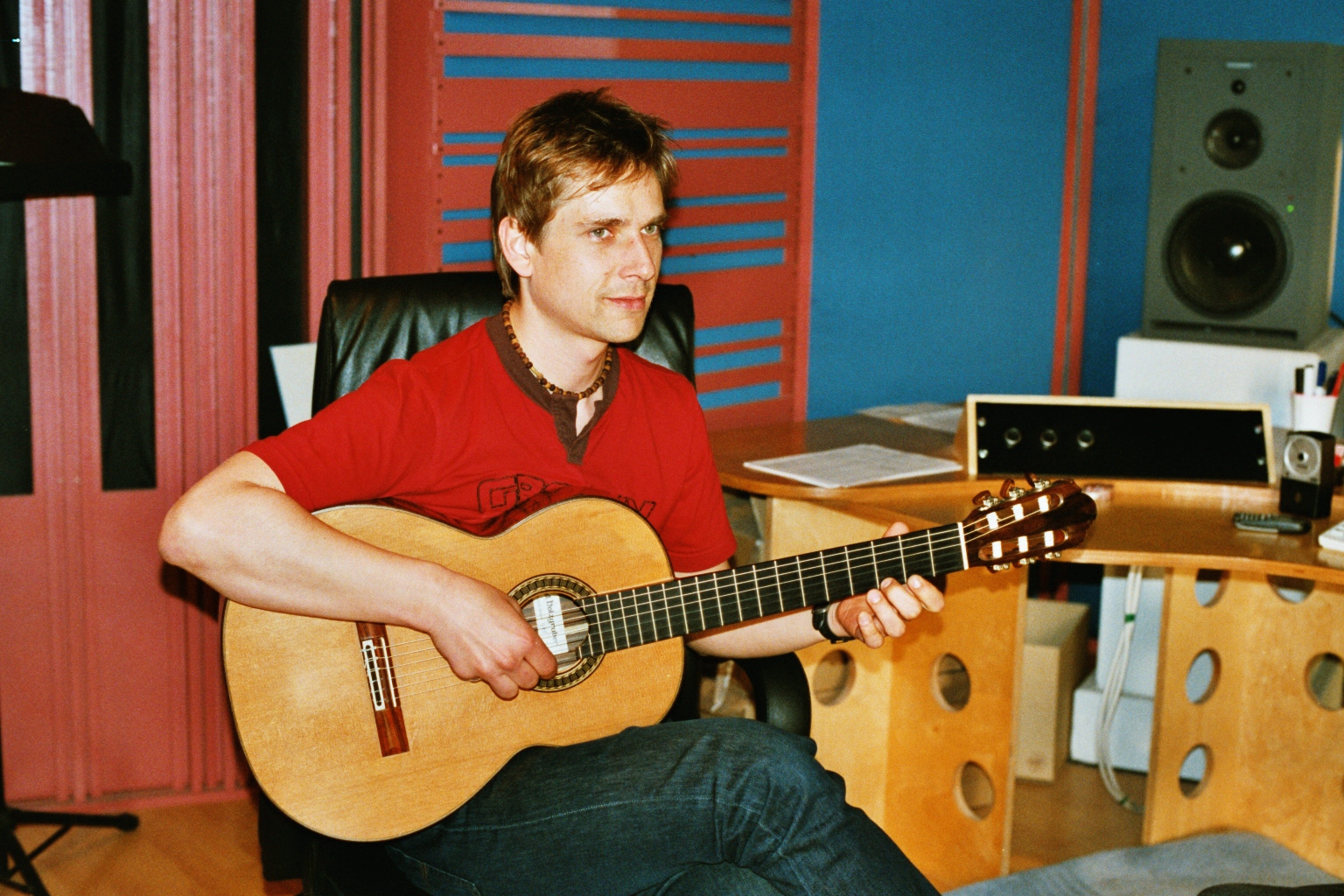 Musician in a red shirt playing an acoustic guitar in a professional music studio with audio equipment.