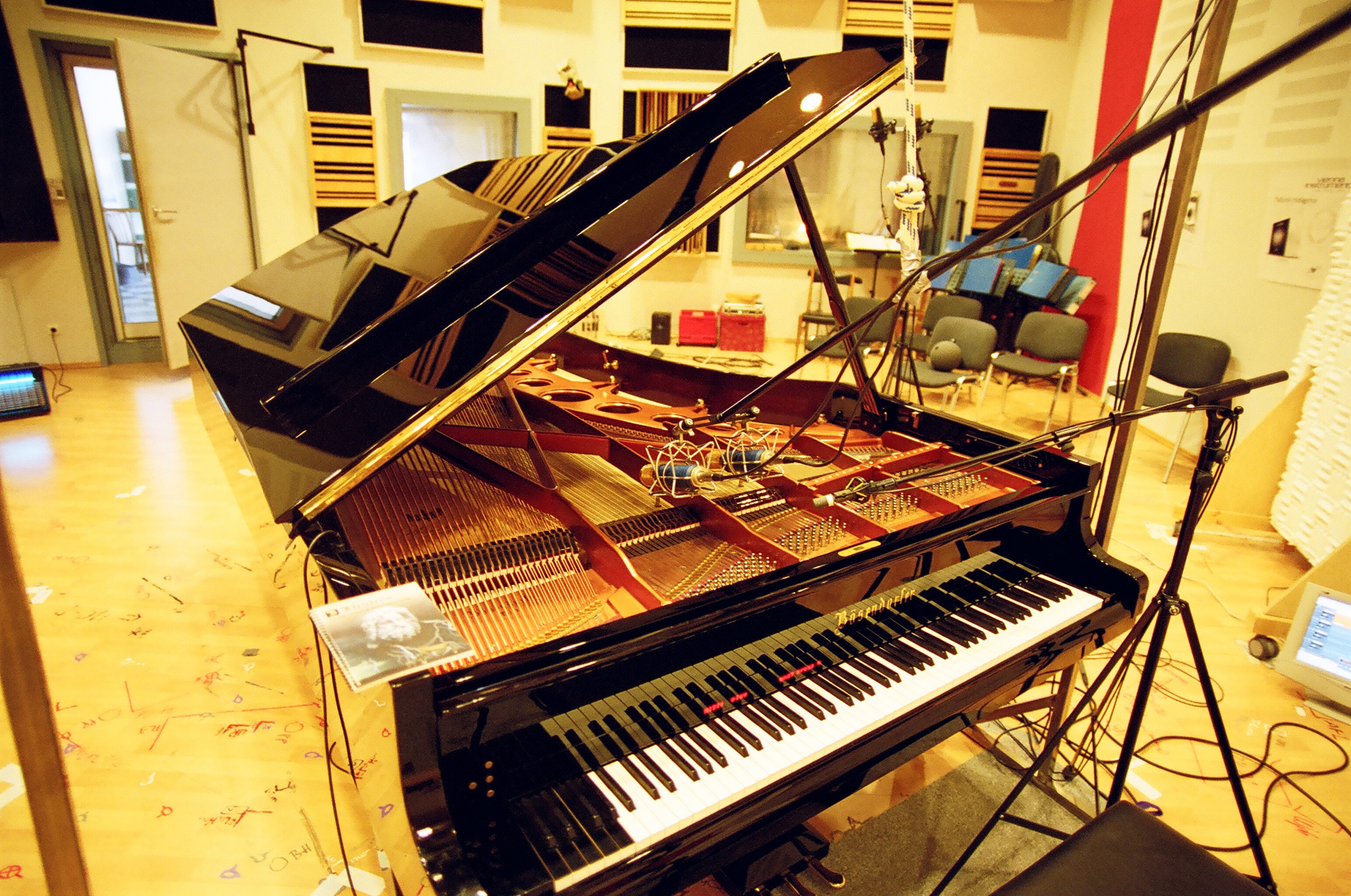 Bösendorfer grand piano with lid open, revealing internal strings and microphones, positioned in a recording studio with sound-dampening panels on walls and a light wooden floor.