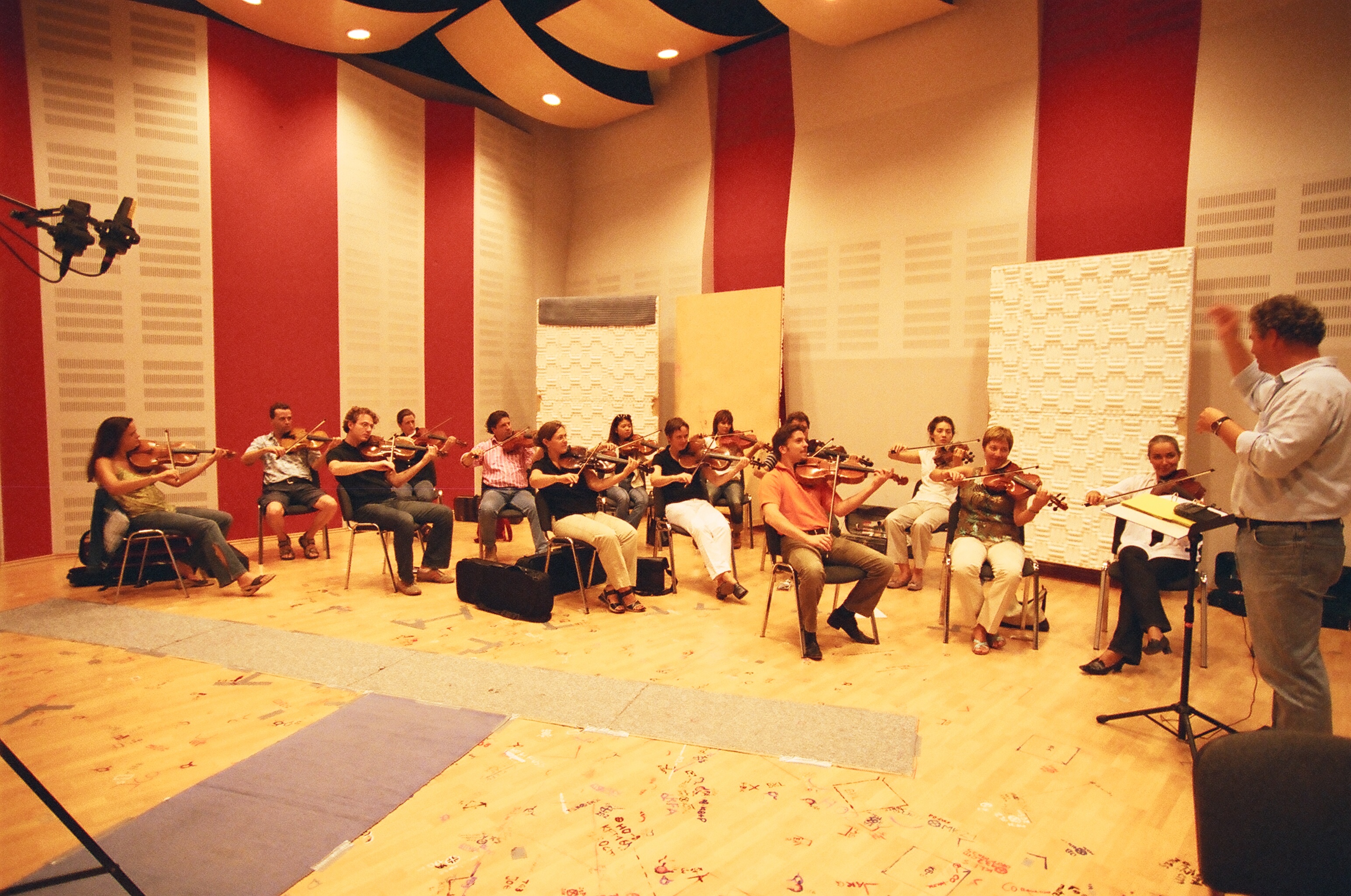 Black and white image of a string orchestra playing violins in a recording studio, with a conductor leading them.