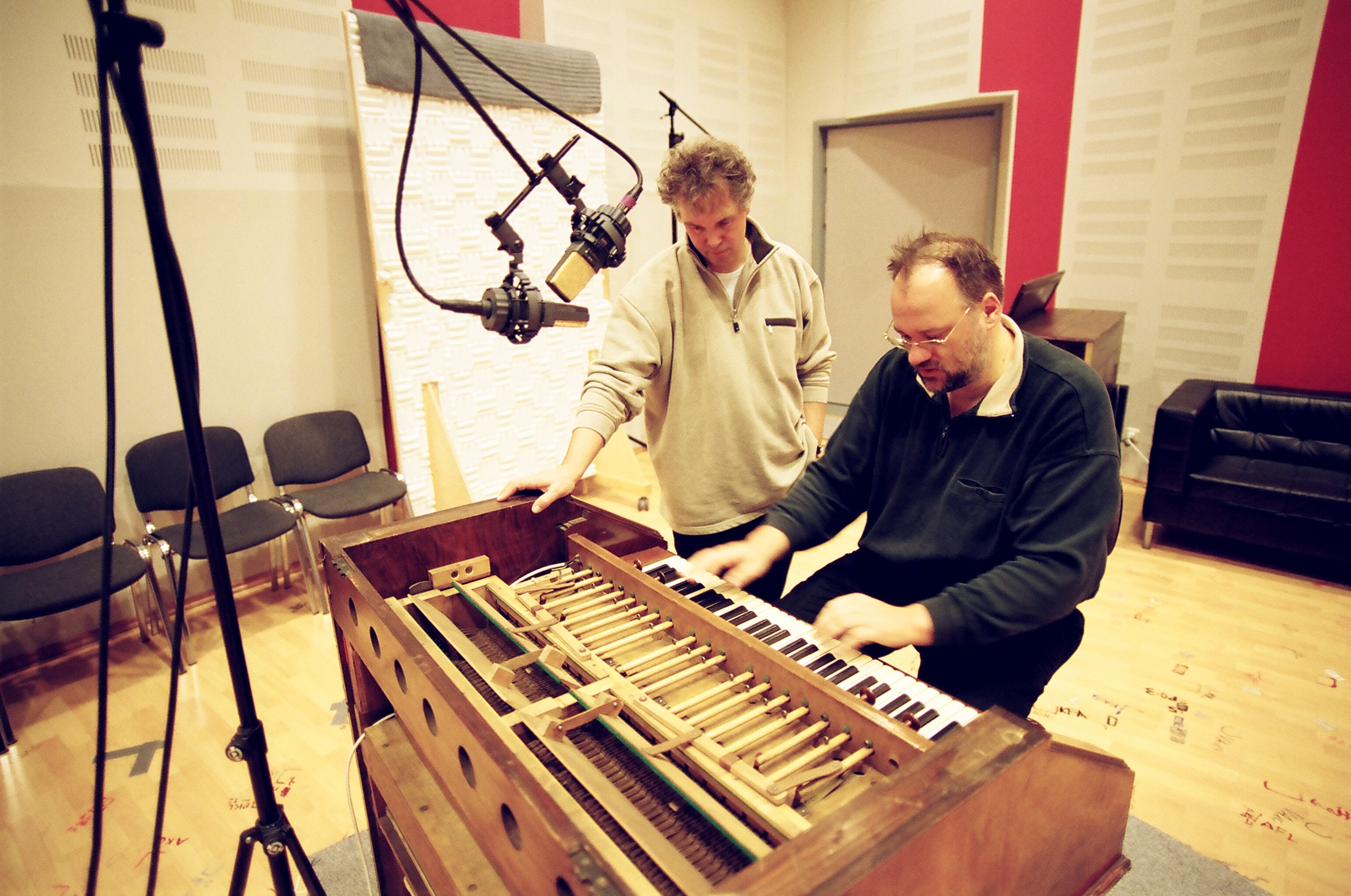 Two men in a recording studio, one playing an open harpsichord or similar keyboard instrument, while the other observes, with microphones positioned nearby.