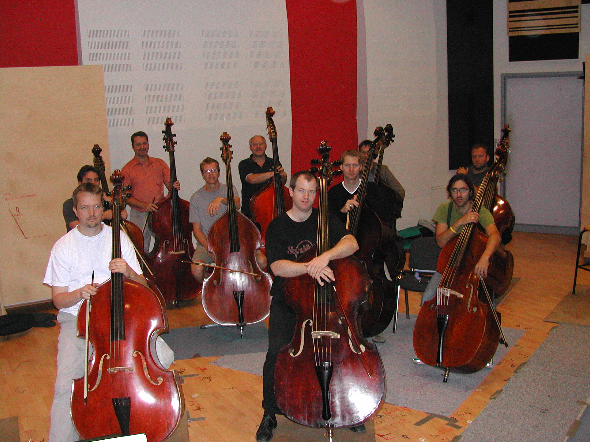 A group of male musicians with multiple dark brown and red double basses, some holding bows, standing in a large room with white and red walls and a light brown floor.