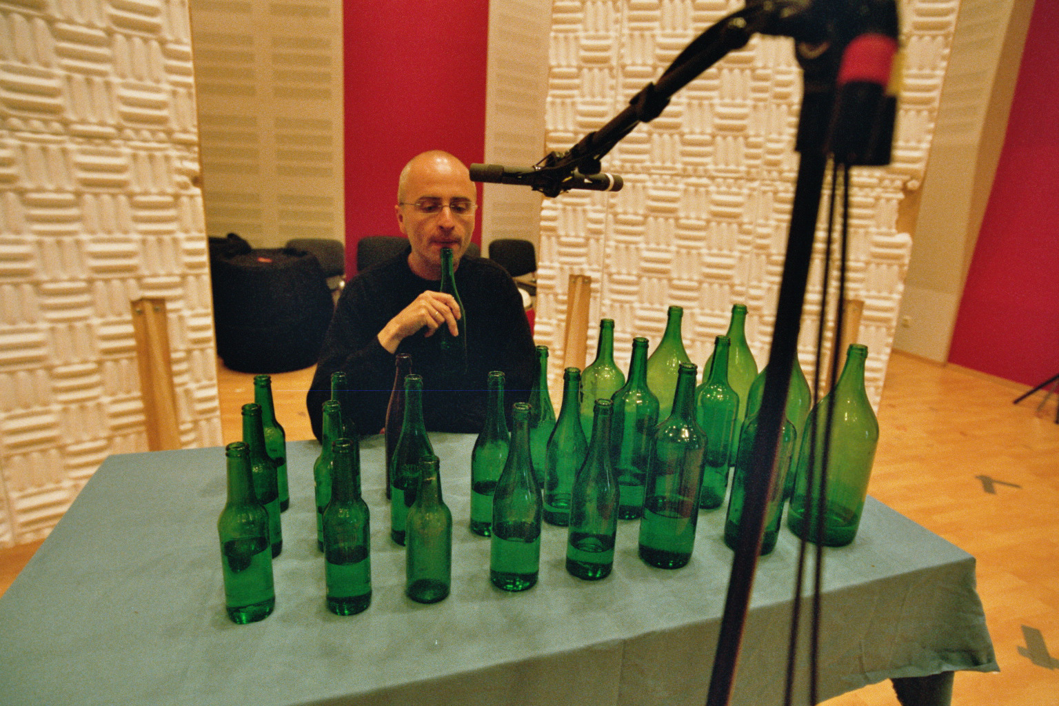 Bald man plays green bottle instrument, surrounded by microphones and soundproofing in a studio, table with many green bottles.