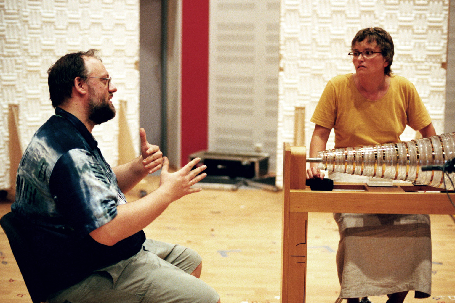 Two musicians in a soundproofed studio, a man seated on the left gesticulating while a woman on the right plays a glass armonica.