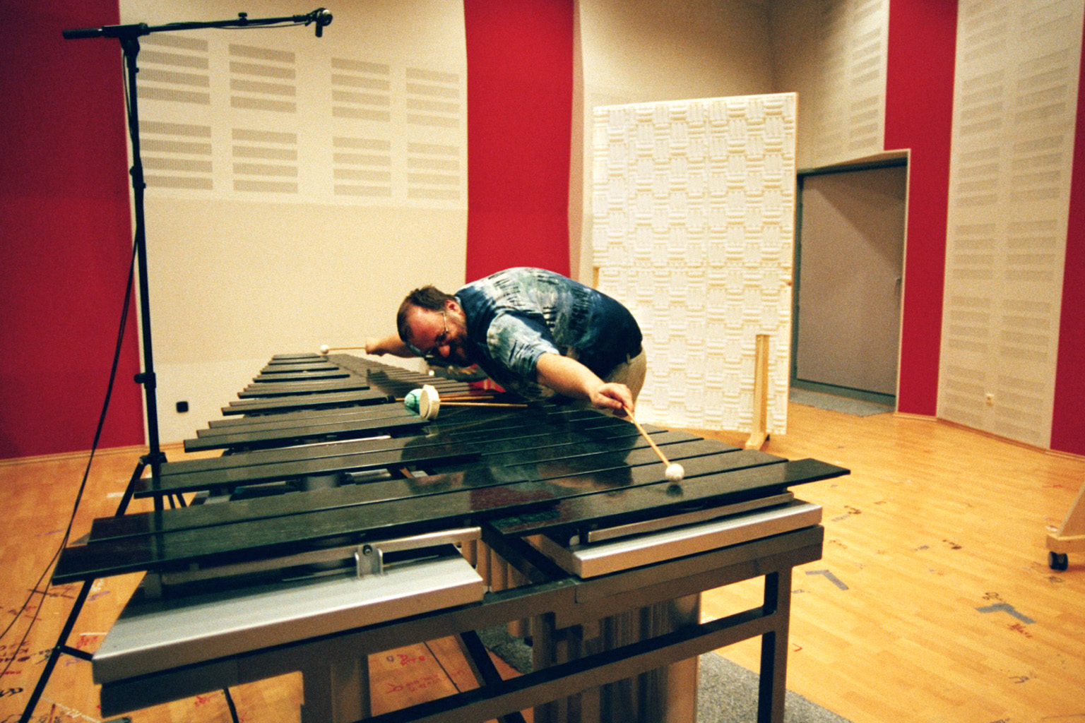 Musician playing a marimba or vibraphone in a recording studio with red and white acoustic panels and a microphone stand.