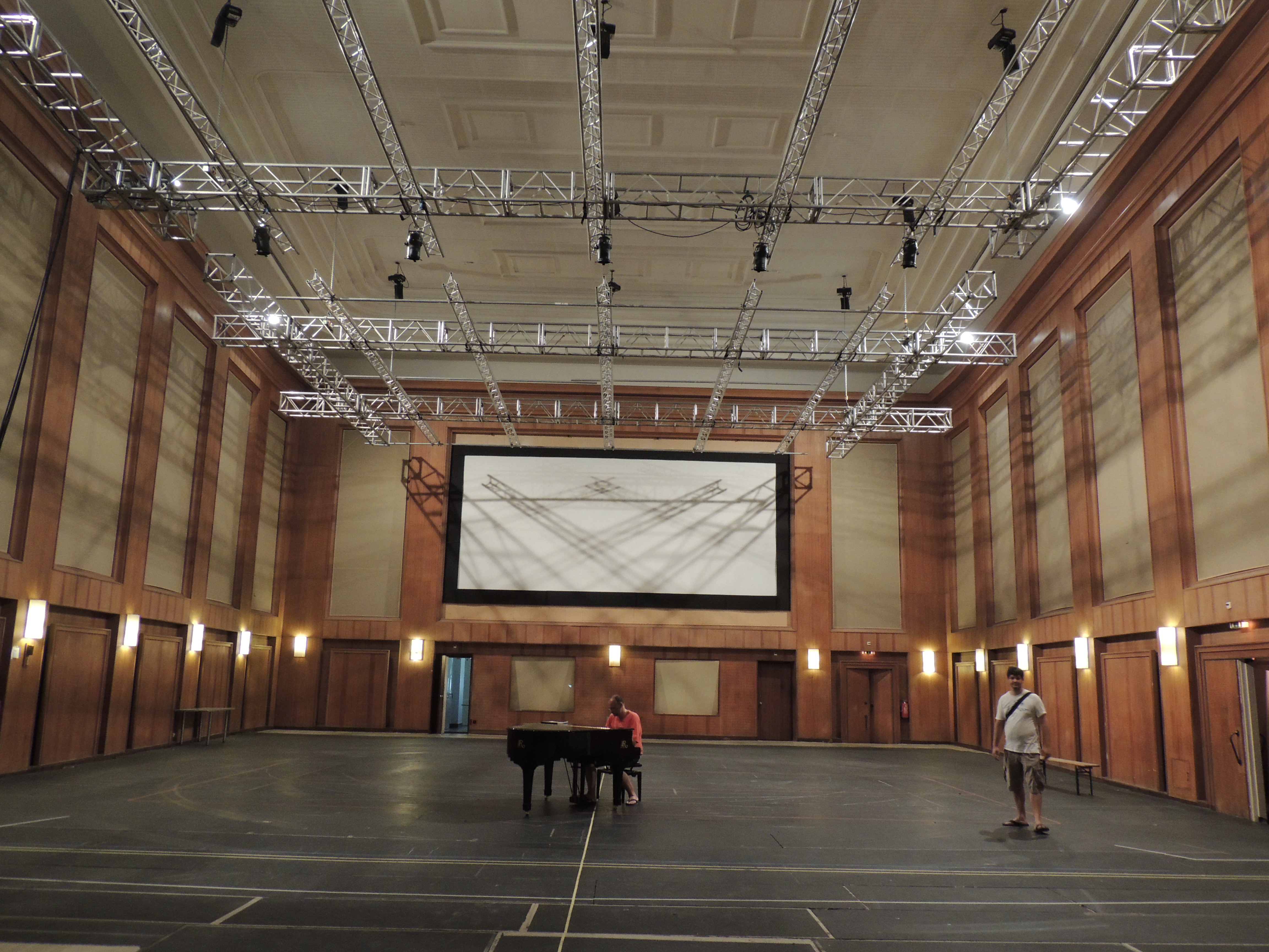 Large empty hall with wooden wall panels, a raised white screen, metal lighting trusses on the ceiling, a person playing a grand piano, and another person standing nearby.