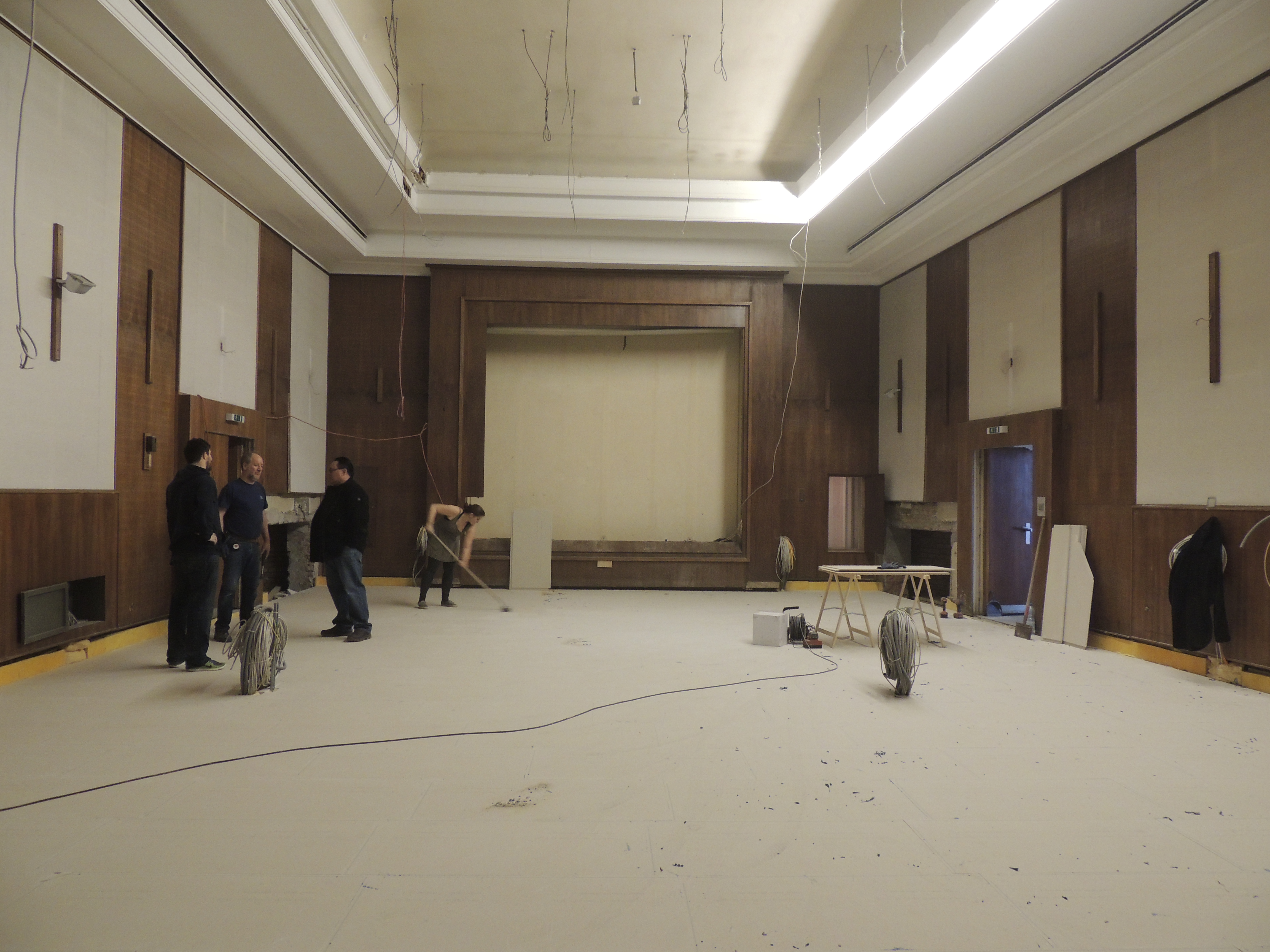 Renovation in progress of a large room with wood paneling, featuring workers, construction materials, and a stage area.