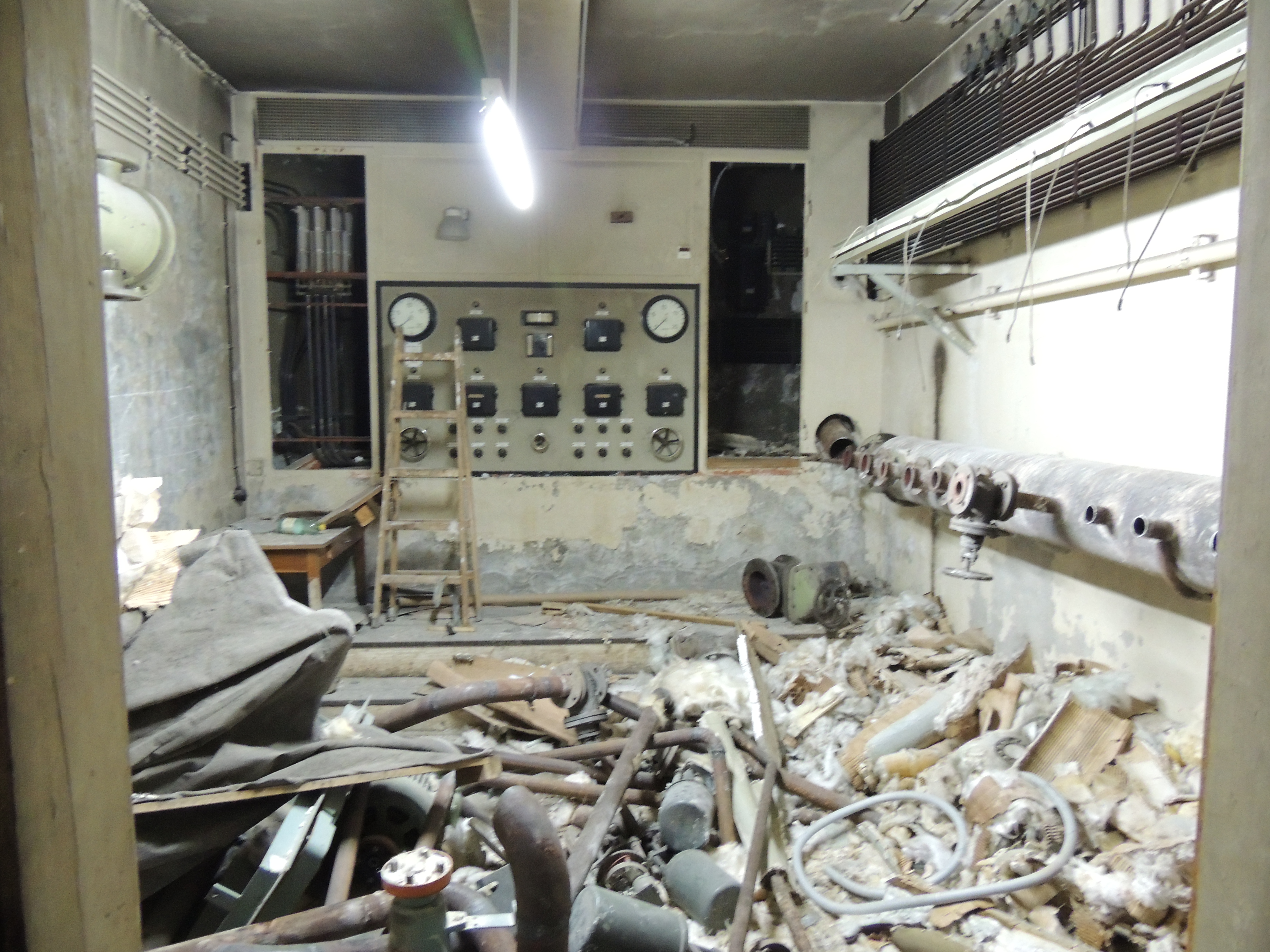 An abandoned, dilapidated industrial control room with a large panel of gauges and switches, a wooden ladder, exposed pipes, and debris scattered across the floor, suggesting disuse and decay.