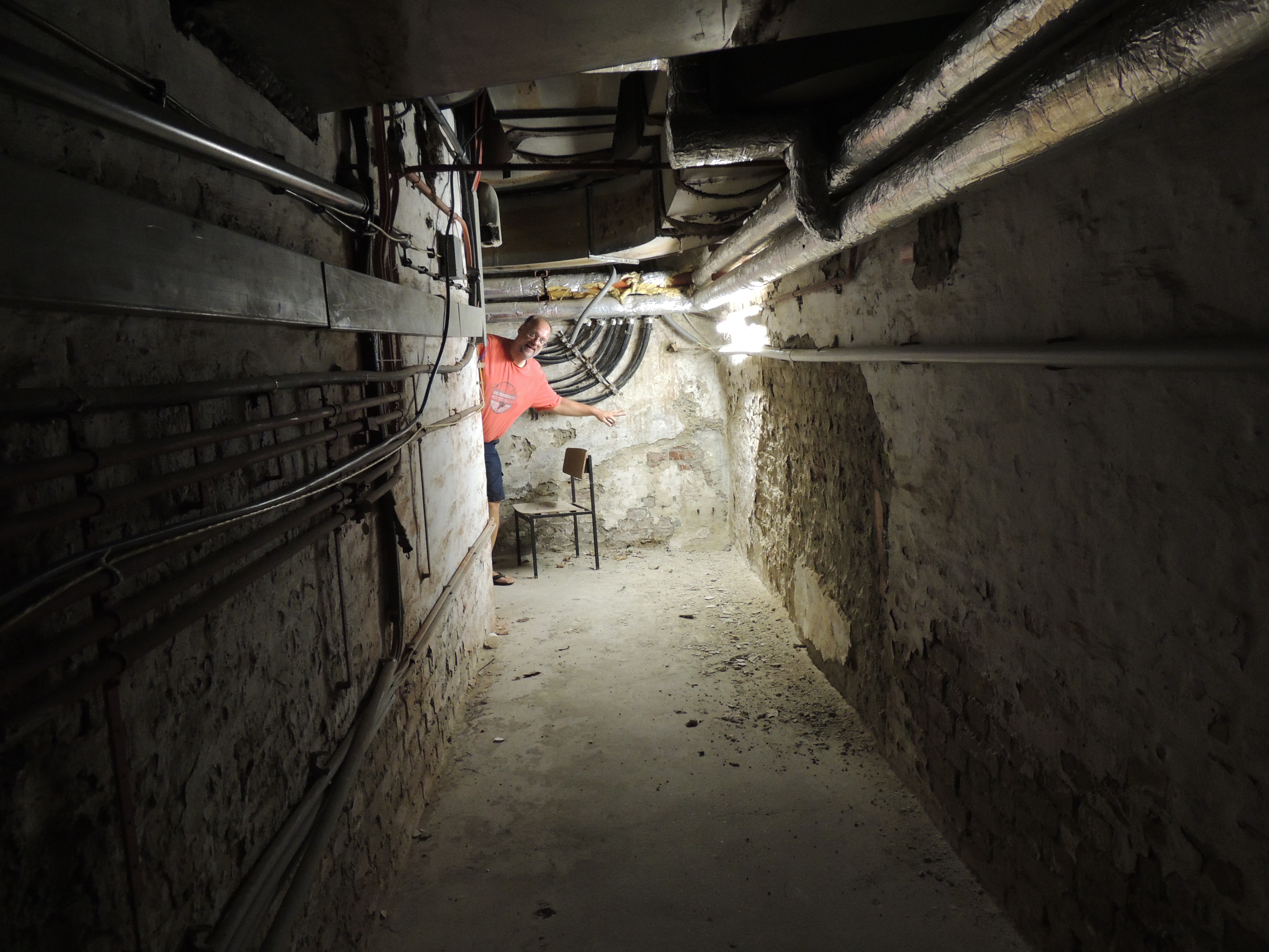 A man points into a dark, narrow, concrete-walled basement corridor, filled with numerous exposed pipes, ducts, and electrical conduits.