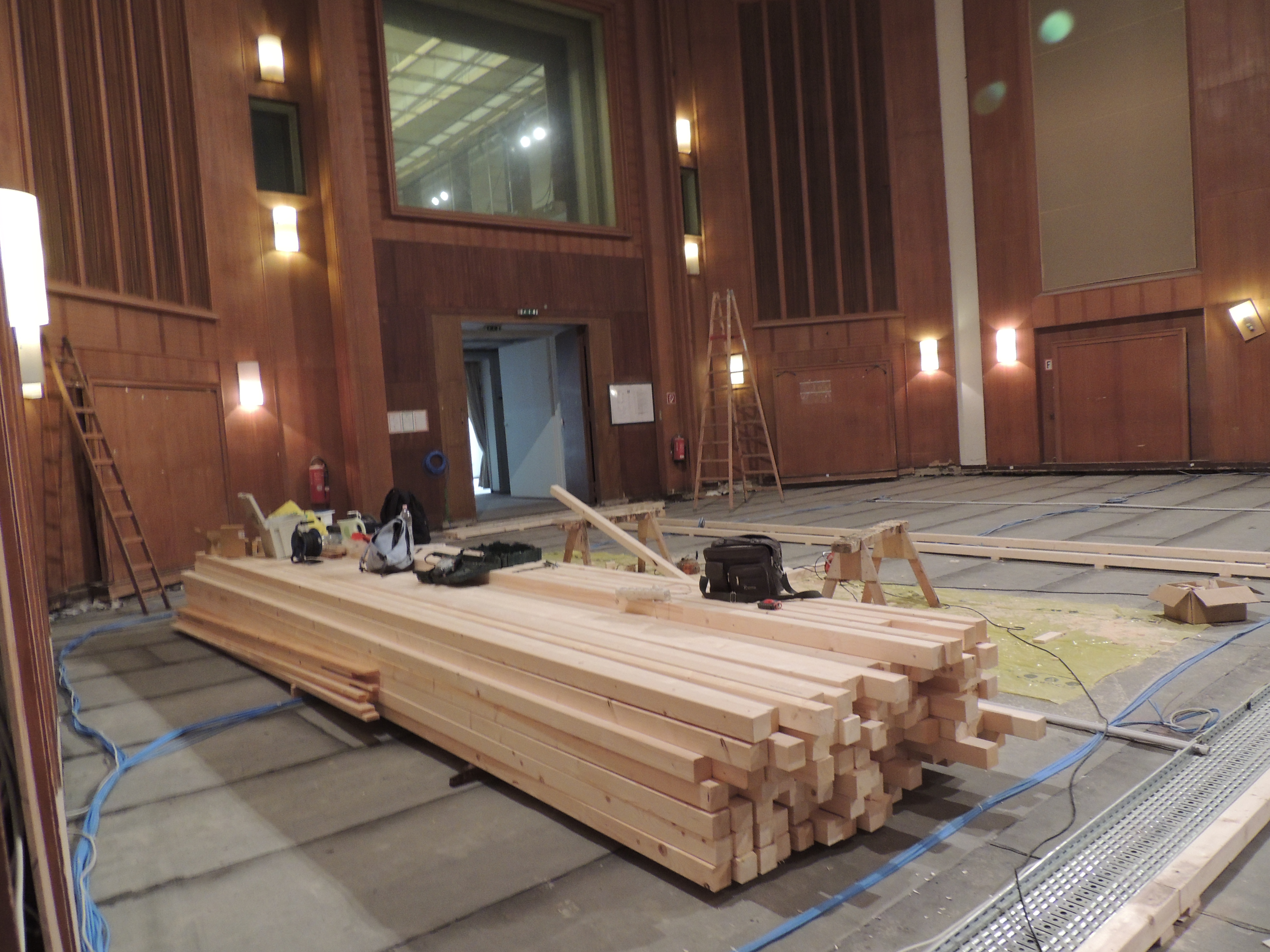 Lumber and construction materials on the floor of a large meeting hall with wood paneled walls, preparing for renovation.