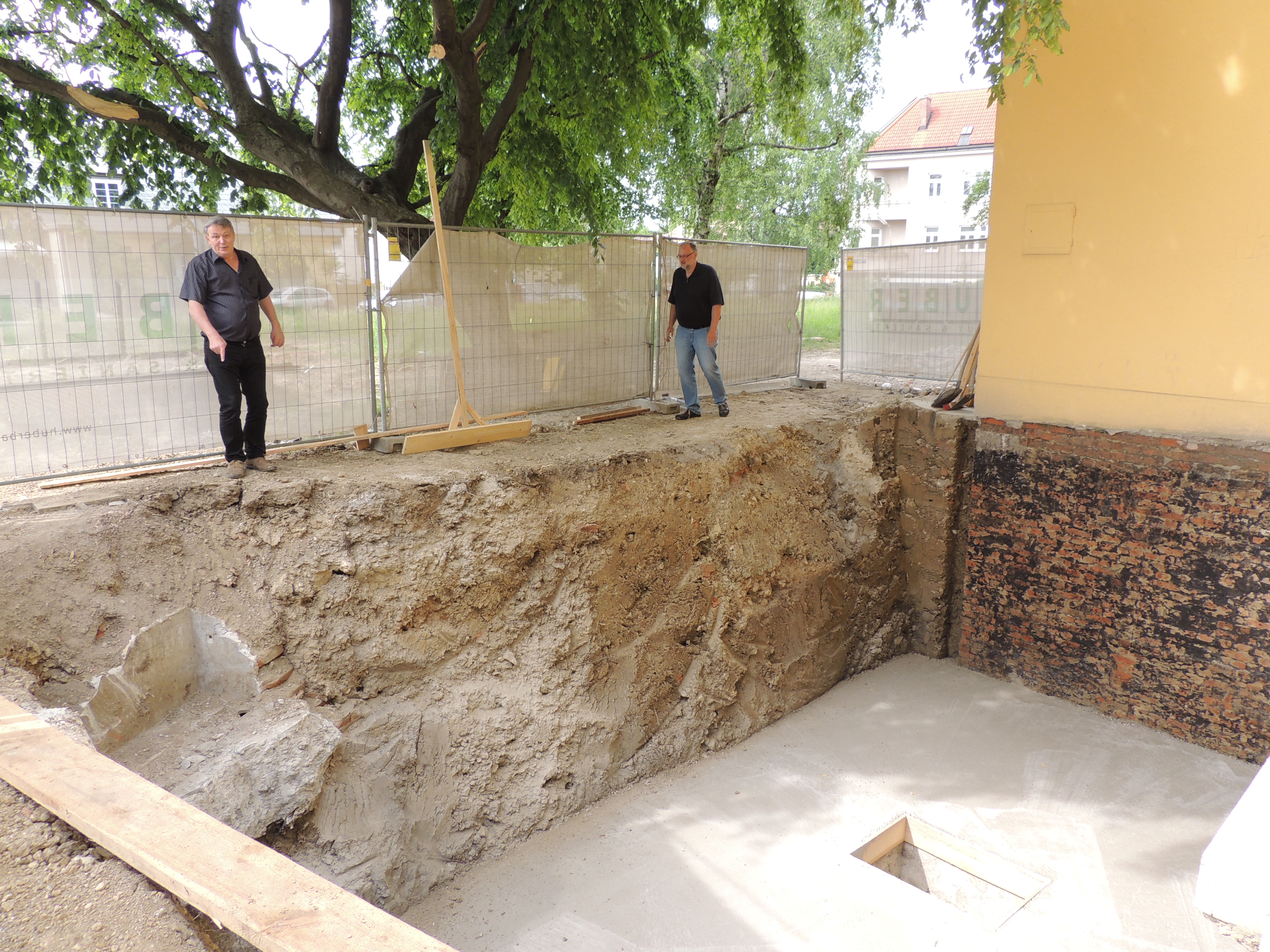 Construction site excavation with two men observing, a concrete foundation laid, and exposed brick walls, featuring a large tree and temporary fencing in the background.
