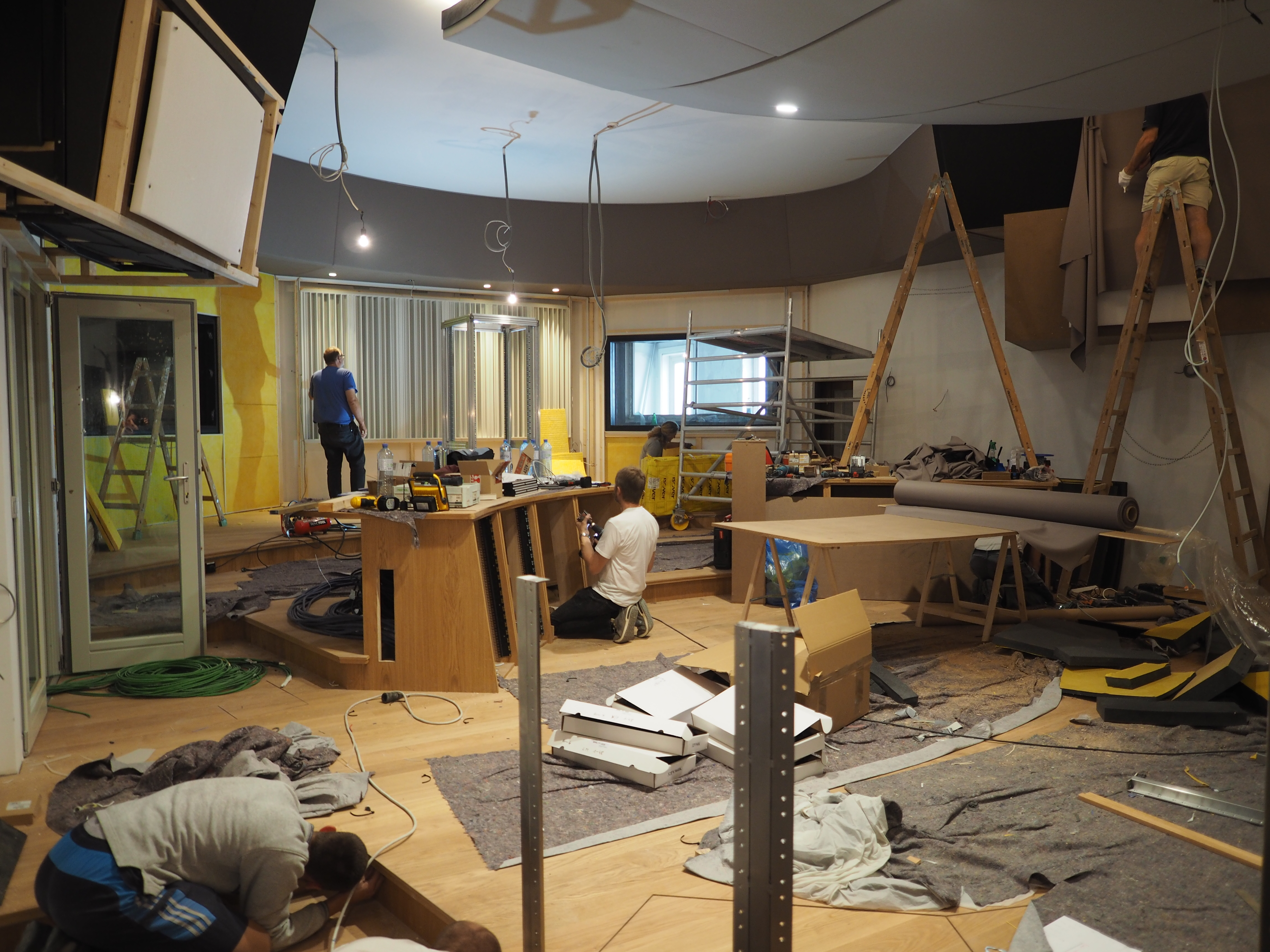 Multiple people install soundproofing and equipment in a large recording studio space with wooden flooring and various construction materials.
