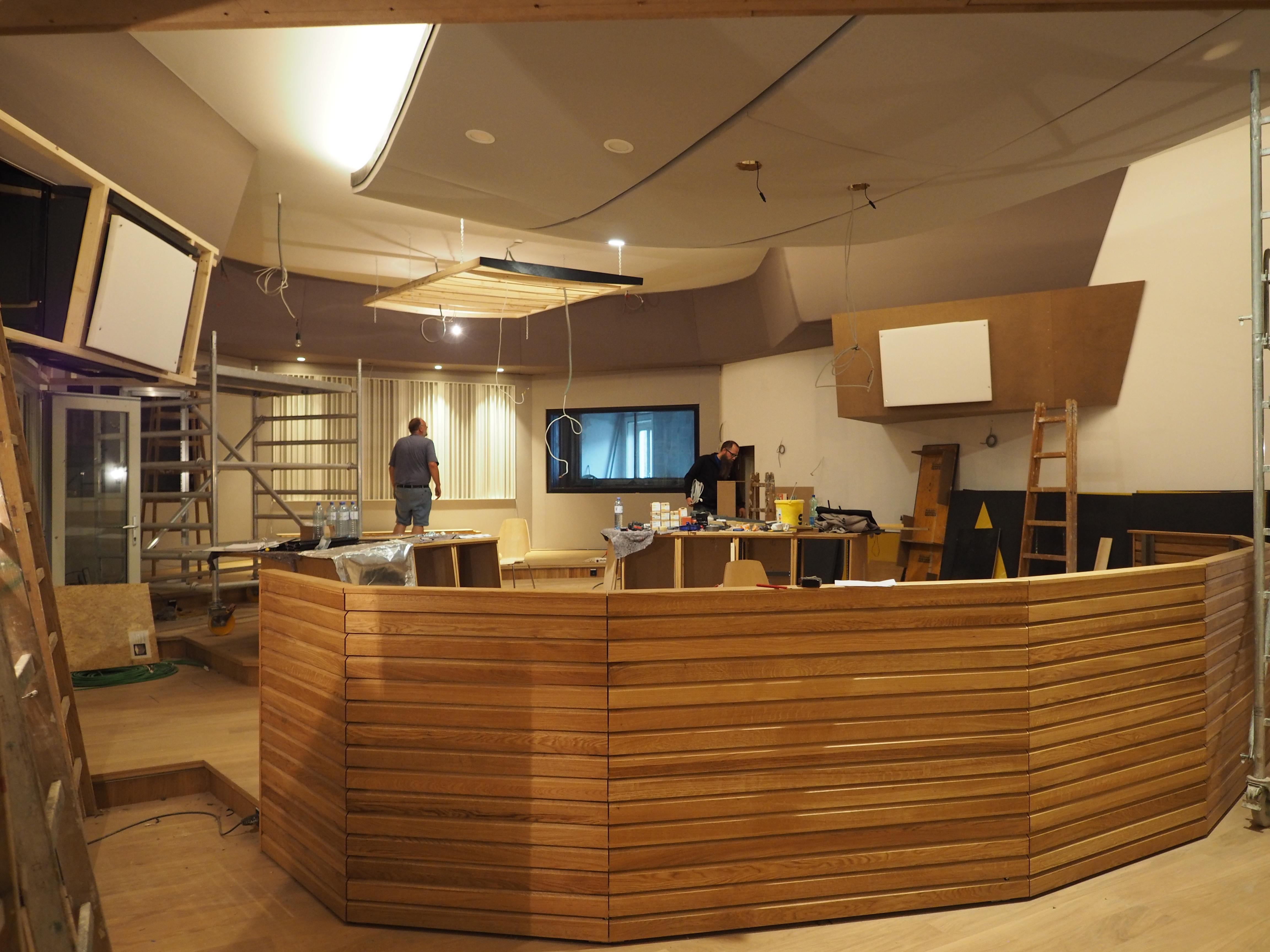 Interior view of a modern studio under construction, featuring a curved wooden counter, ceiling-mounted sound panels, and workers installing equipment.