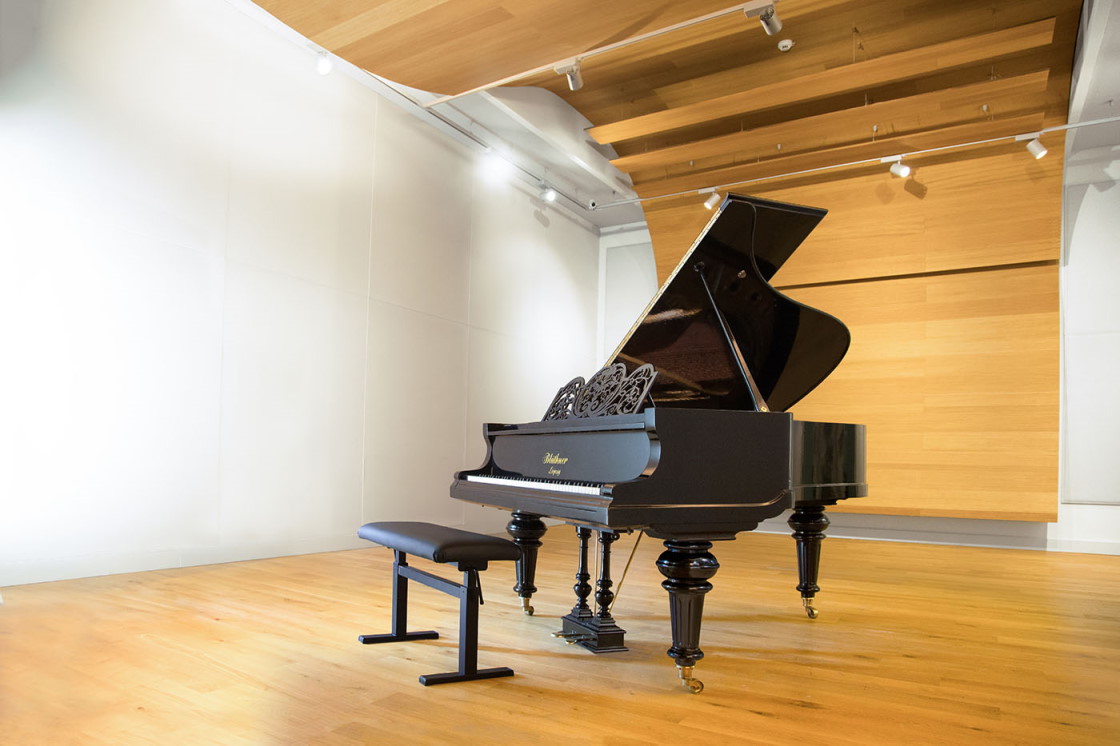 Glossy black Blüthner grand piano, built in 1895, with a dark bench on a light wood floor in a modern room with white and wood panelled walls.
