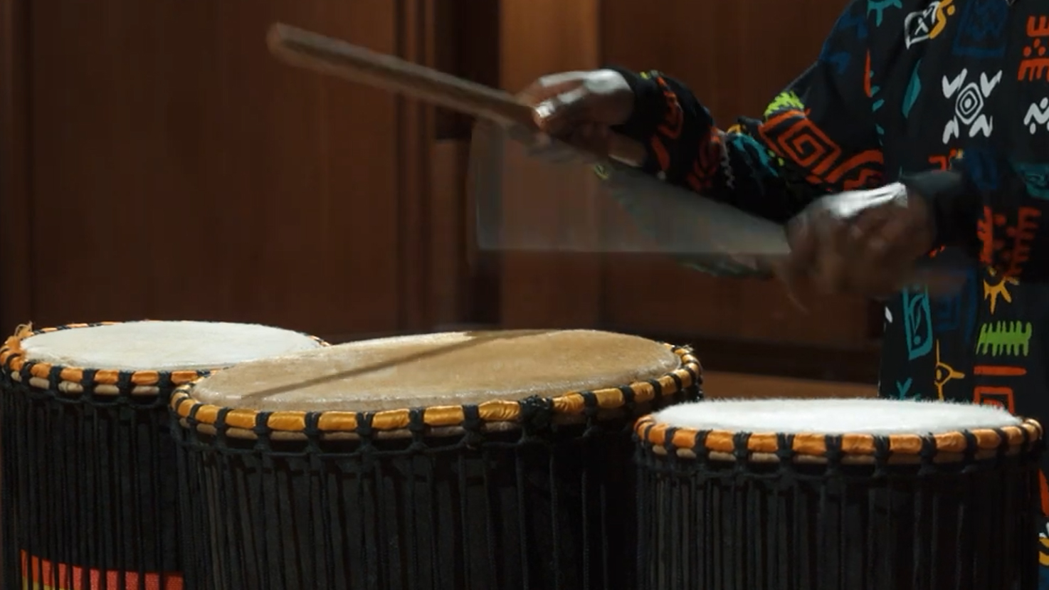 Person drumming with sticks on a set of three djembe drums, wearing a colorful, patterned shirt.