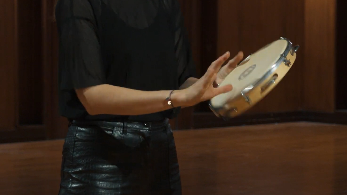 Person playing a tambourine, holding the musical instrument with both hands while wearing a sheer black top and black leather skirt.