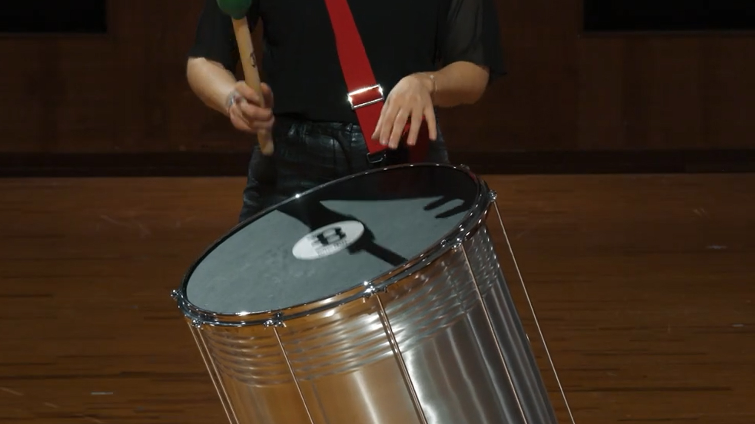 Musician playing a drum with a green mallet, wearing black clothing and red suspenders.