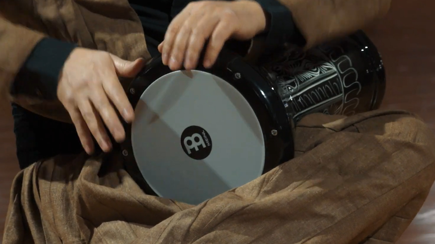 Close-up of a person's hands playing a Meinl darbuka drum, showcasing the white drumhead and decorative black and silver body, with the player's brown pants visible.