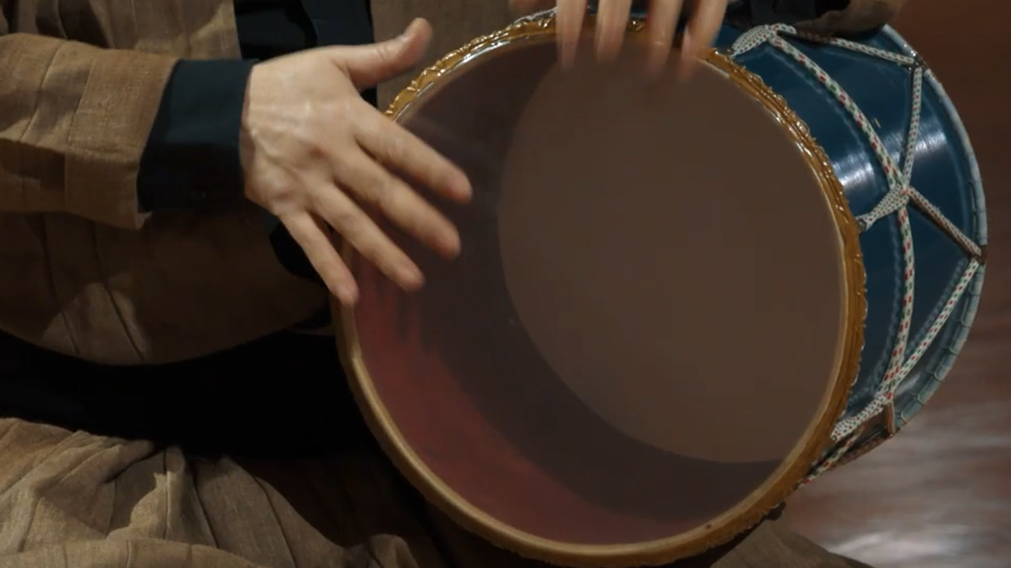 Hands playing a traditional Korean janggu drum, showing its two drumheads and blue lacquered body with woven white rope.