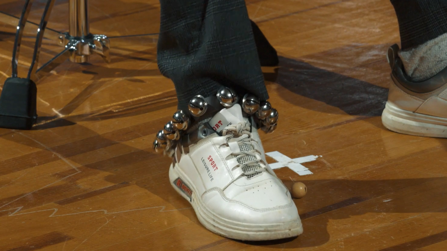 Close-up of a person's foot wearing a white sneaker with silver jingle bells attached around the ankle, standing on a wooden floor, next to a drum pedal.