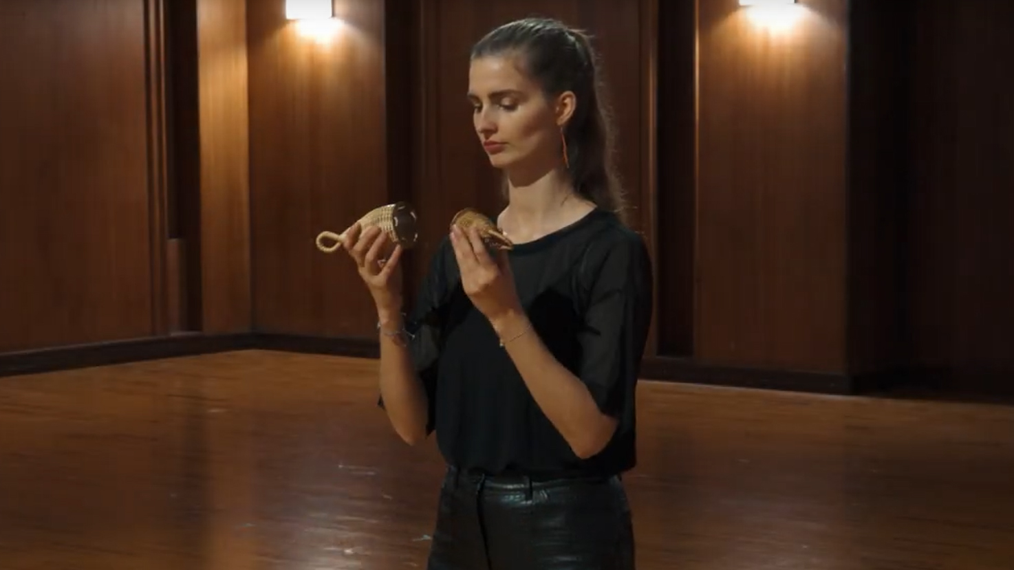 Young woman in black top and dark pants holding two woven rattles or shakers, standing on a wooden floor against a paneled wall with lights.