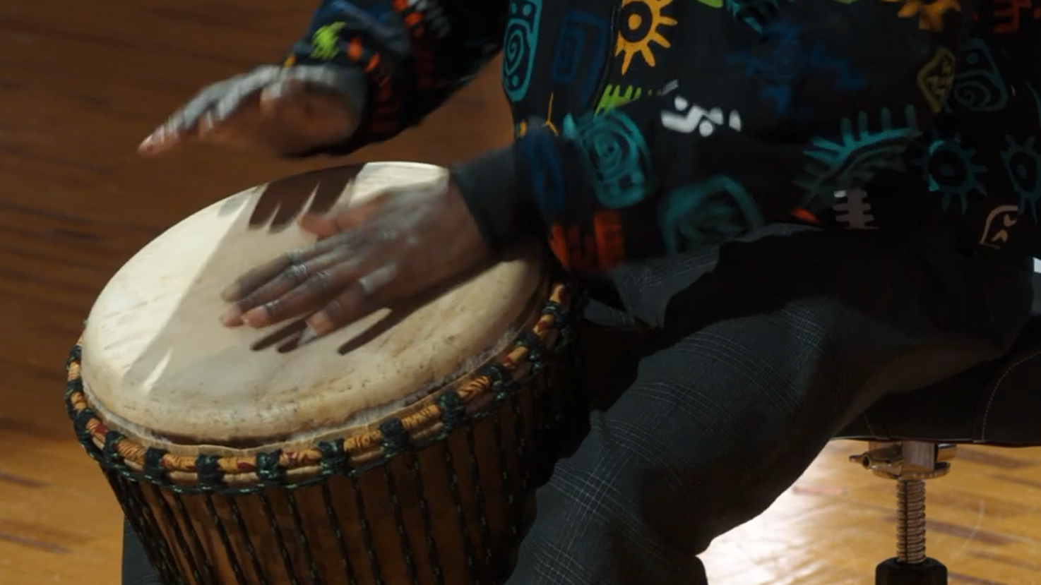 Person wearing colorful tribal print shirt playing a djembe drum, hands blurred from motion.