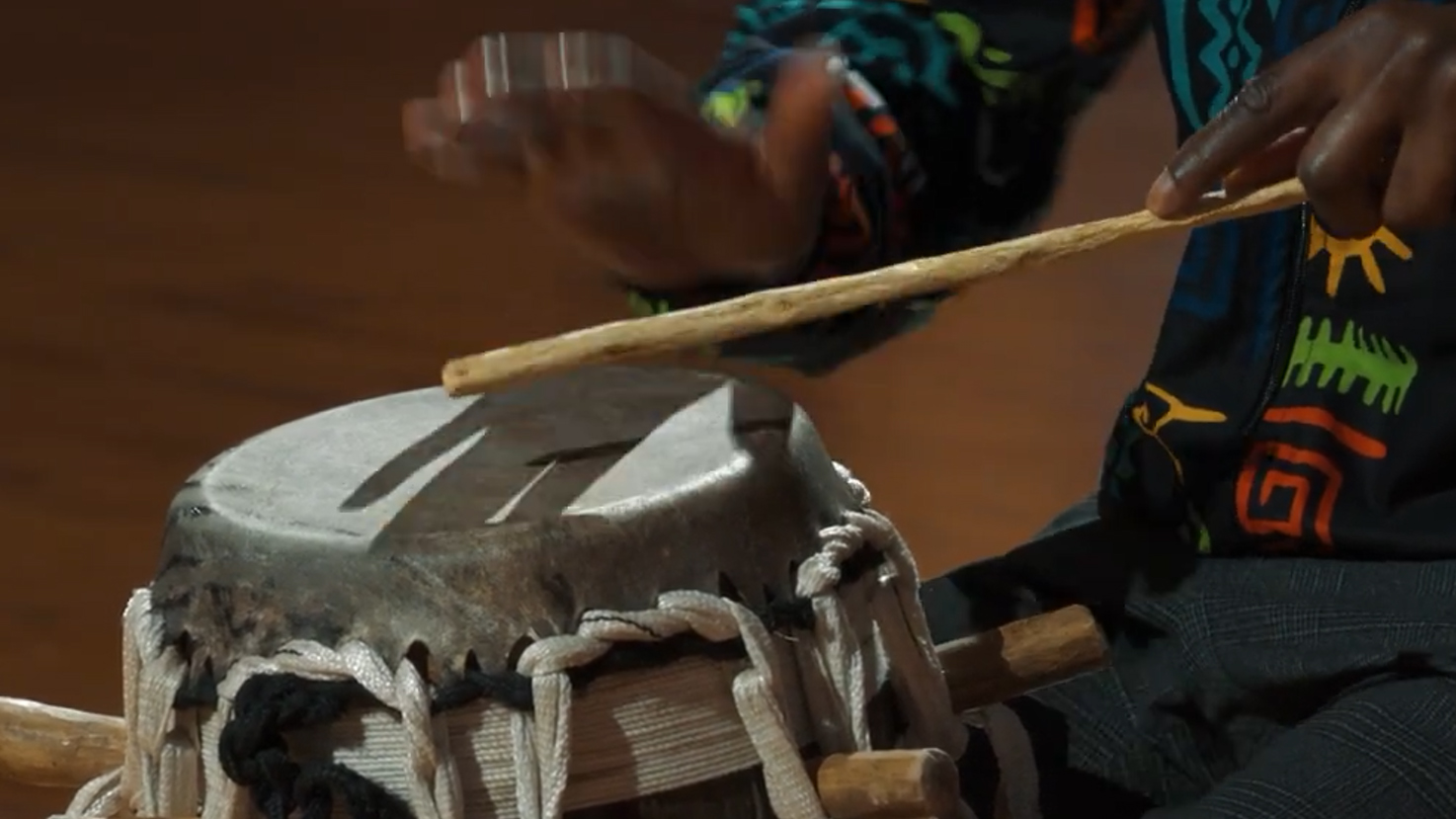 Person playing a djembe drum with a stick, showcasing African music and traditional instruments.