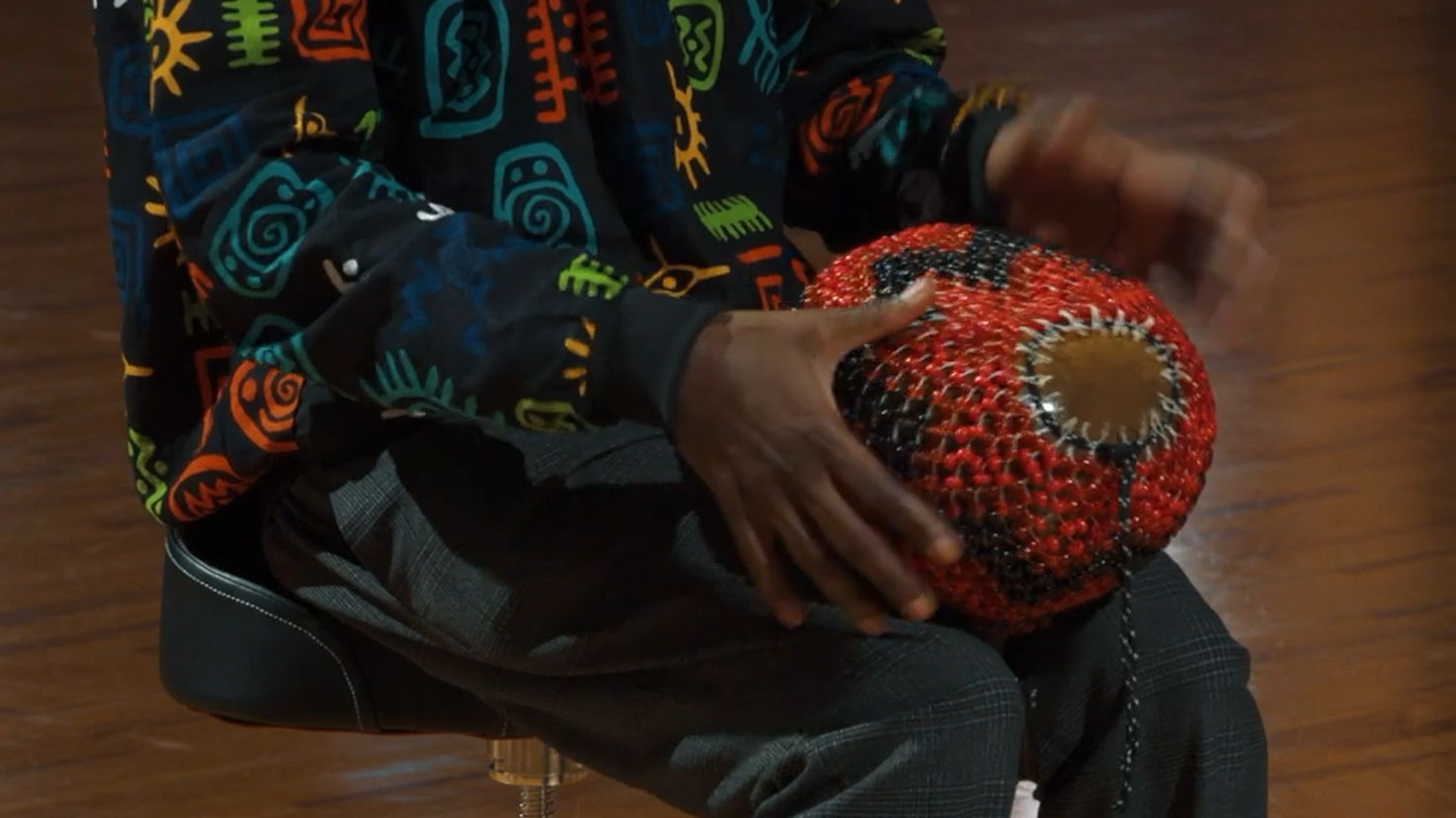 Person in colorful patterned shirt playing a red and black beaded percussion instrument.