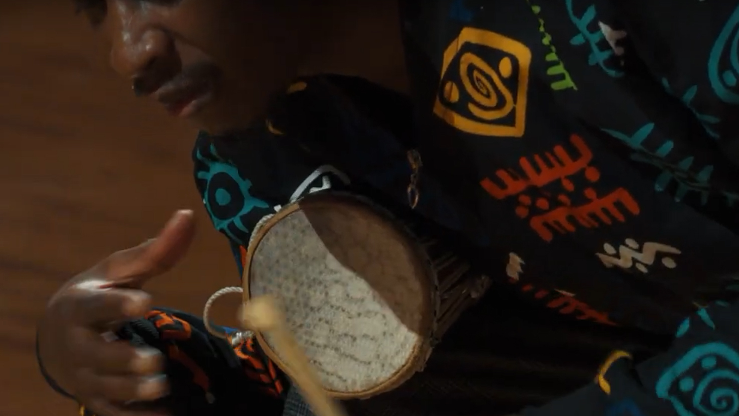 Close-up of a person wearing clothing with colorful patterns, playing a hand drum or djembe with a stick.