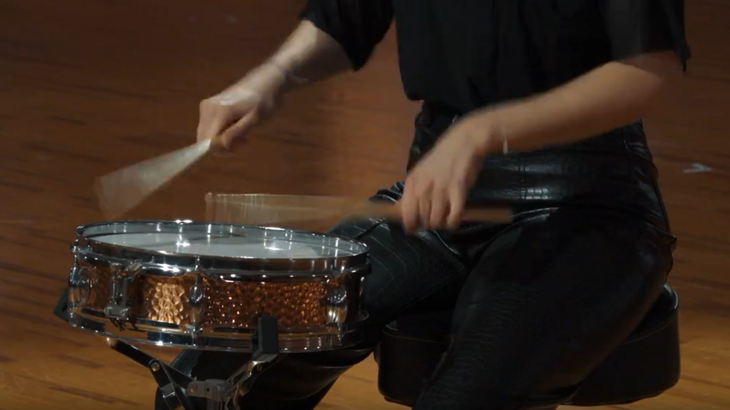 Musician playing a shiny copper snare drum with drumsticks, wearing a black shirt and black leather pants, on a wooden floor.