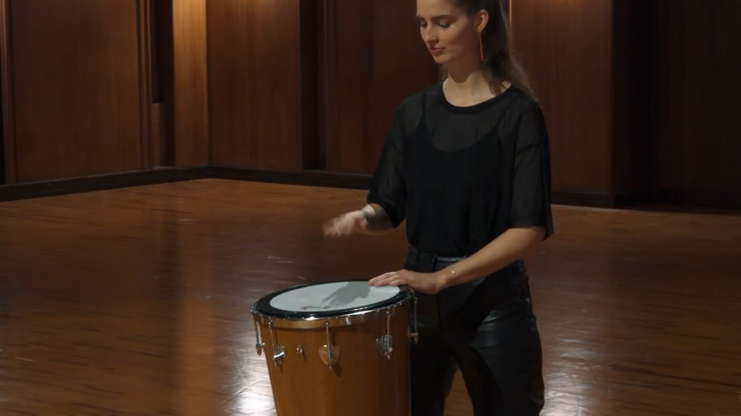 Young woman in a black shirt and leather pants playing a tall drum with both hands in a warm wooden room.
