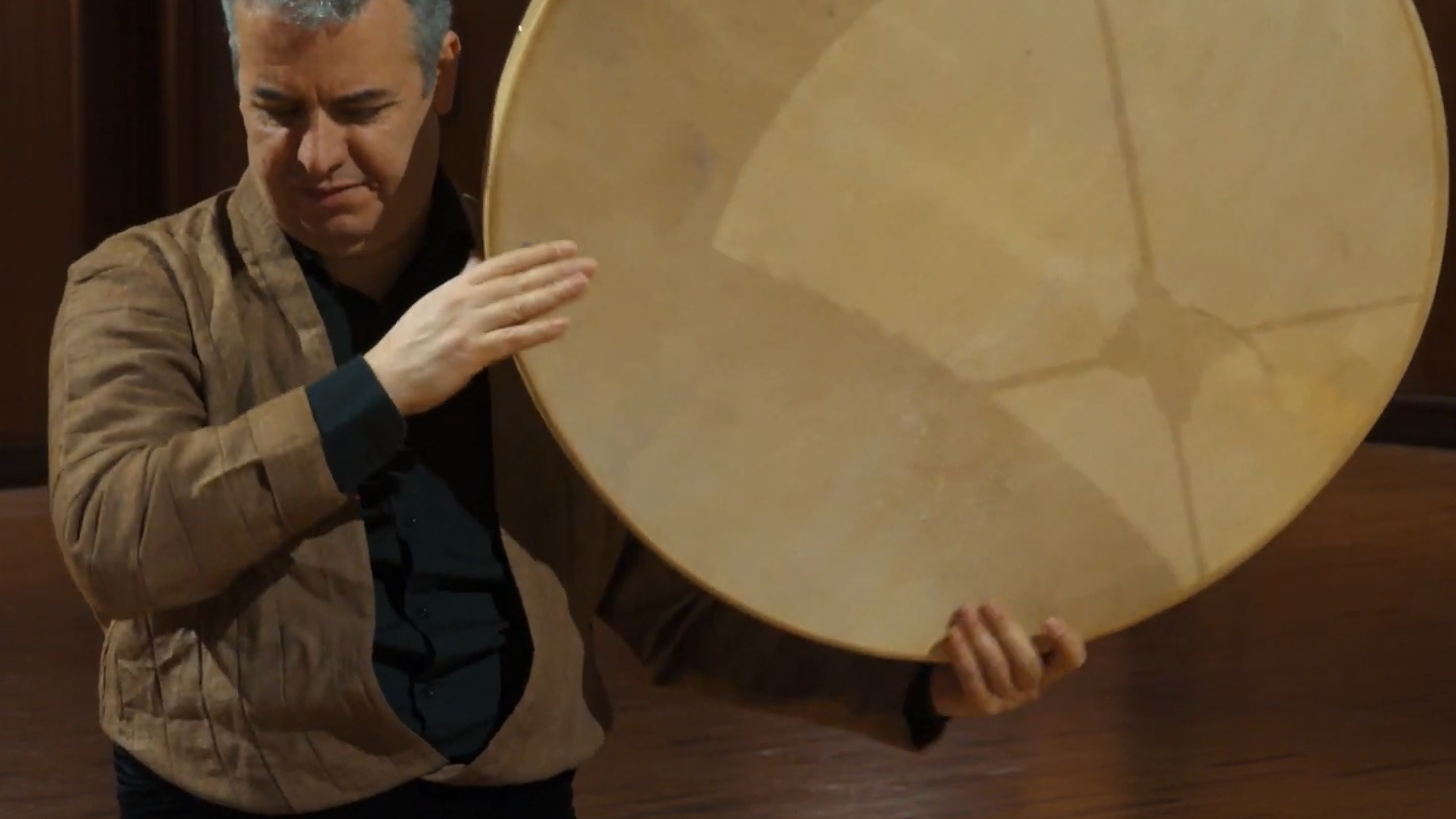 Man playing a frame drum or tambourine, with hands on the drumhead of the musical instrument.