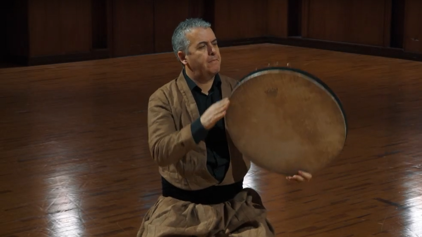 Man in traditional attire playing a hand drum on a wooden floor, showcasing musical performance and cultural instruments.