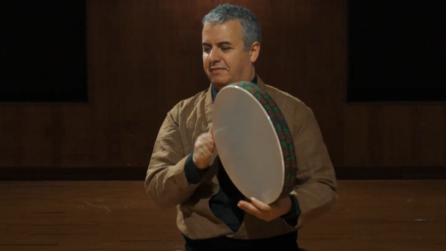 Man playing a frame drum or tambourine, wearing a light brown jacket, performing traditional or world music.