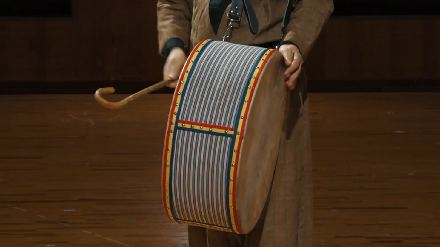 Person in historical clothing playing a colorful striped bass drum with a hooked drumstick, suspended by a shoulder strap against a wooden floor background.