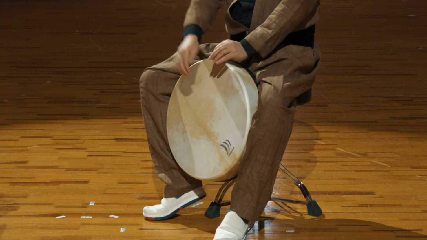 Musician playing a frame drum or bodhrán on a stand, seated on a wooden floor.