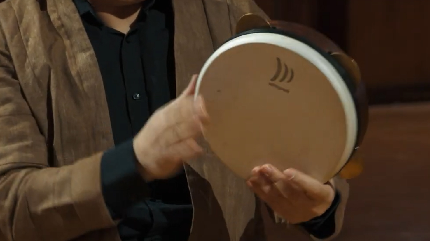 Musician in a brown jacket and black shirt playing a tambourine or frame drum with both hands.