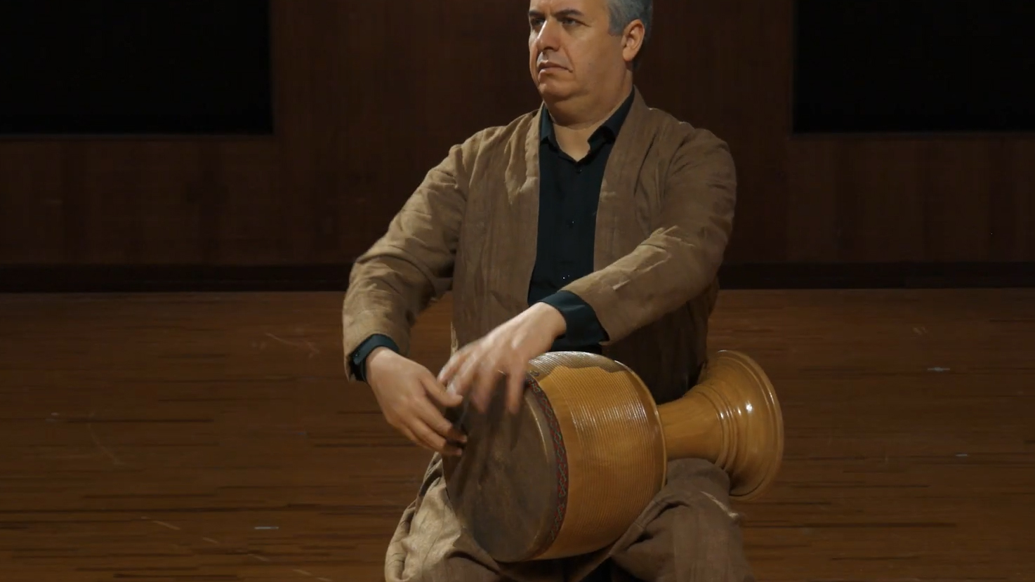 Man playing a tombak, a Persian goblet drum, with both hands, wearing a traditional brown tunic and black shirt, focused during a cultural performance or musical recording.