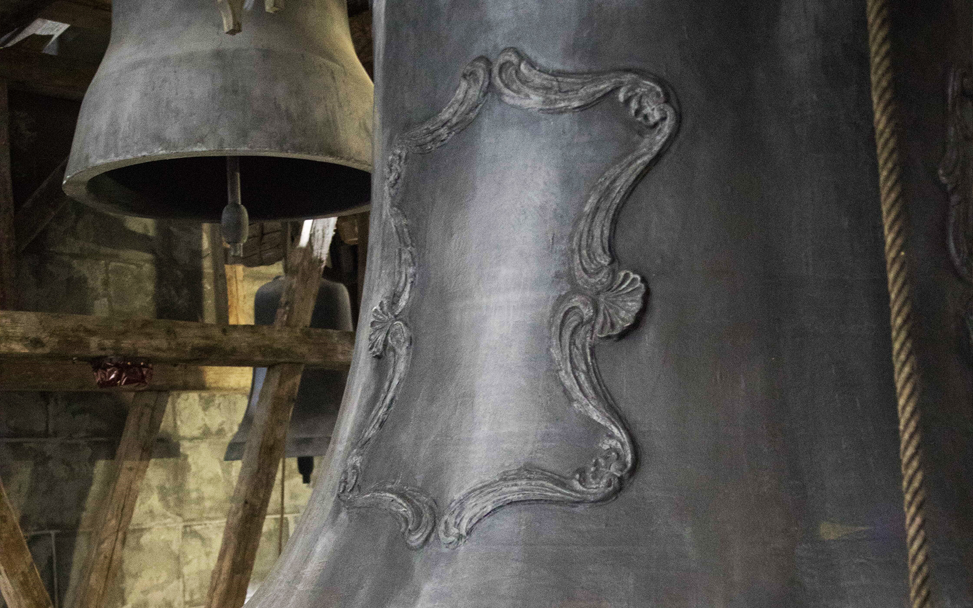 Close-up of a large, dark gray church bell with ornate baroque-style raised detailing, showing the bell's texture and decorative elements. Another unadorned bell is visible in the background, suspended within the bell tower's wooden beams.