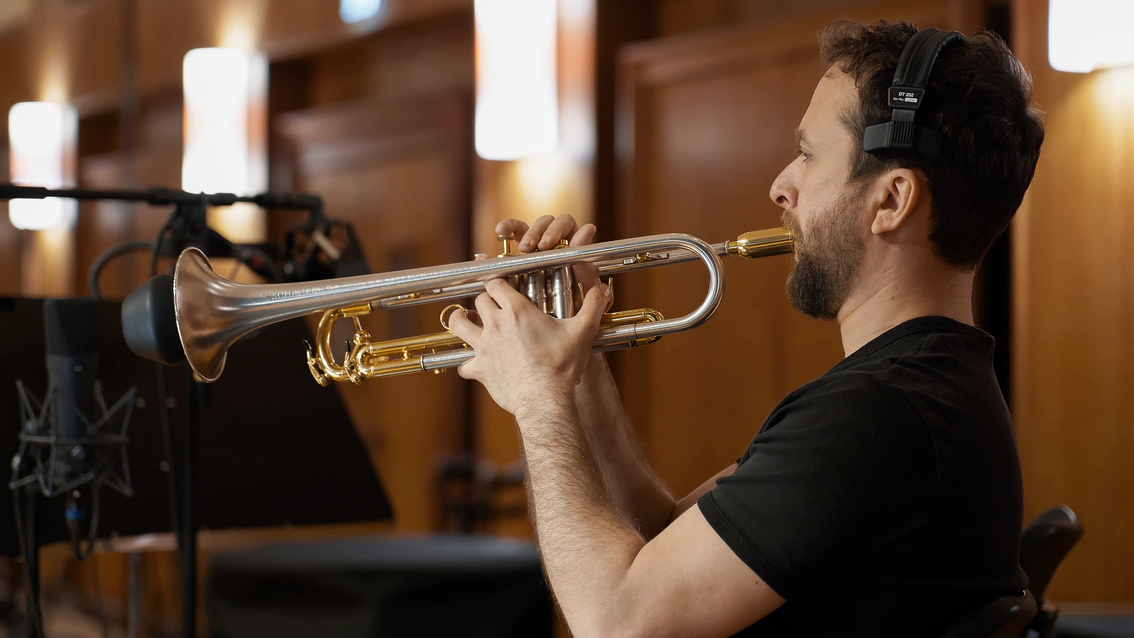 A male musician with a beard and black headphones plays a silver and gold trumpet in a recording studio, with a microphone and light in the background.