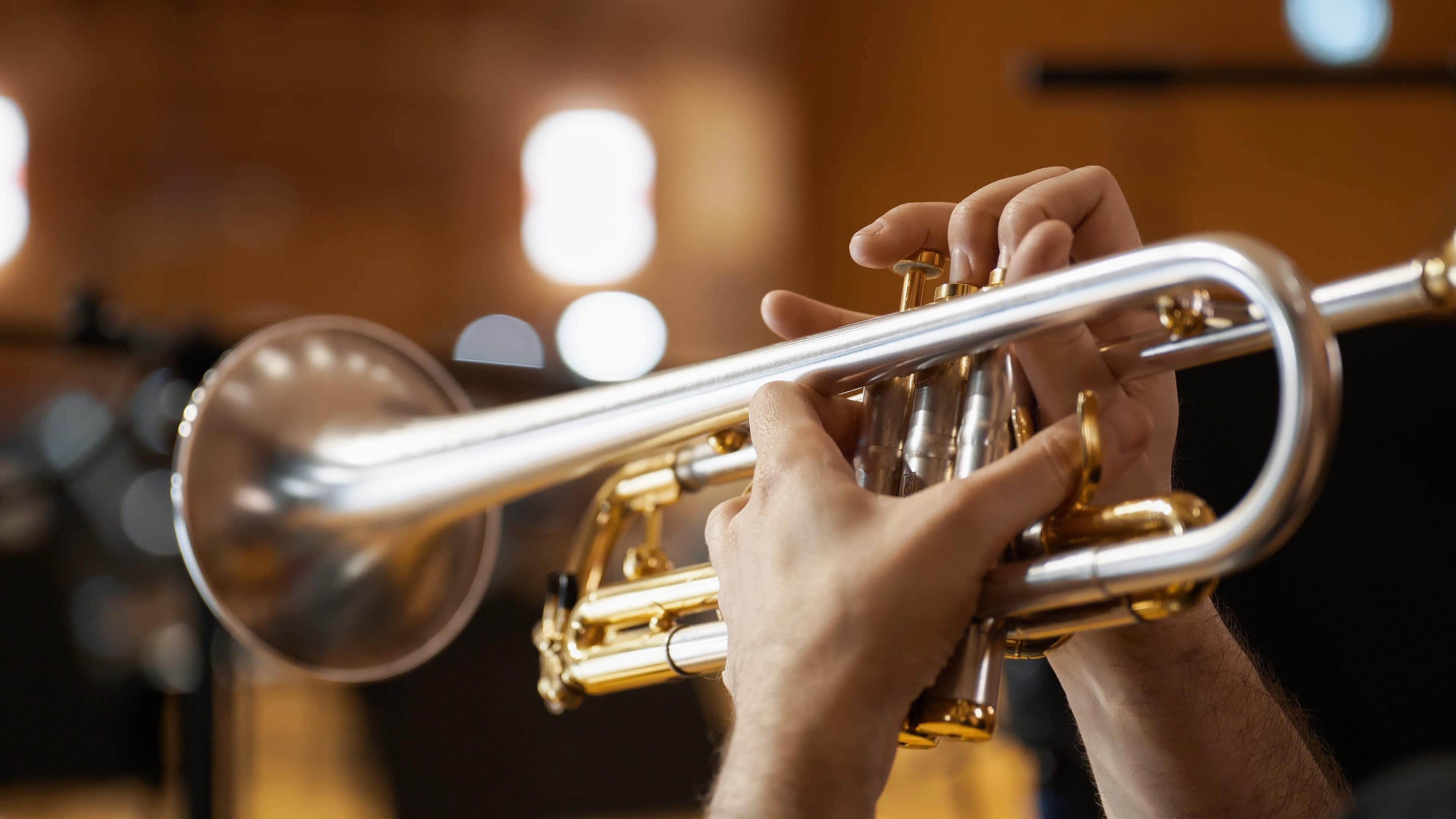 Hands playing a shiny silver and gold trumpet, pressing the valves in a musical performance, with a blurred warm-toned background.