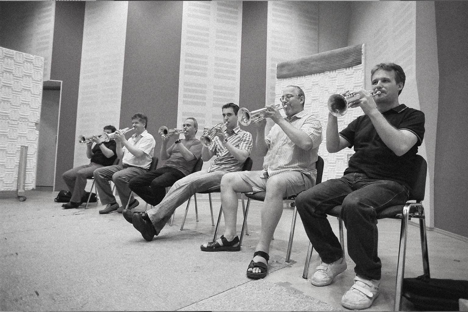 Black and white image of six men sitting in chairs, playing trumpets in what appears to be a rehearsal studio or music room.