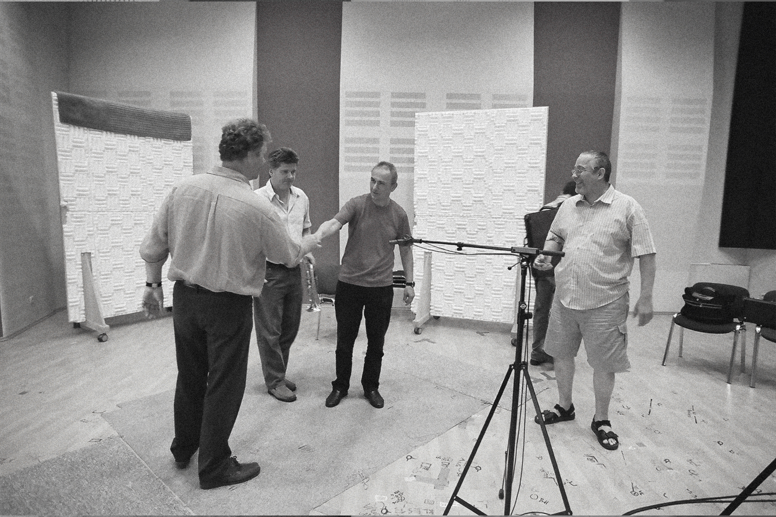 Black and white photo of five men in a large recording studio with soundproofing panels and microphone stands. Two men are shaking hands, while another man holds a trumpet.
