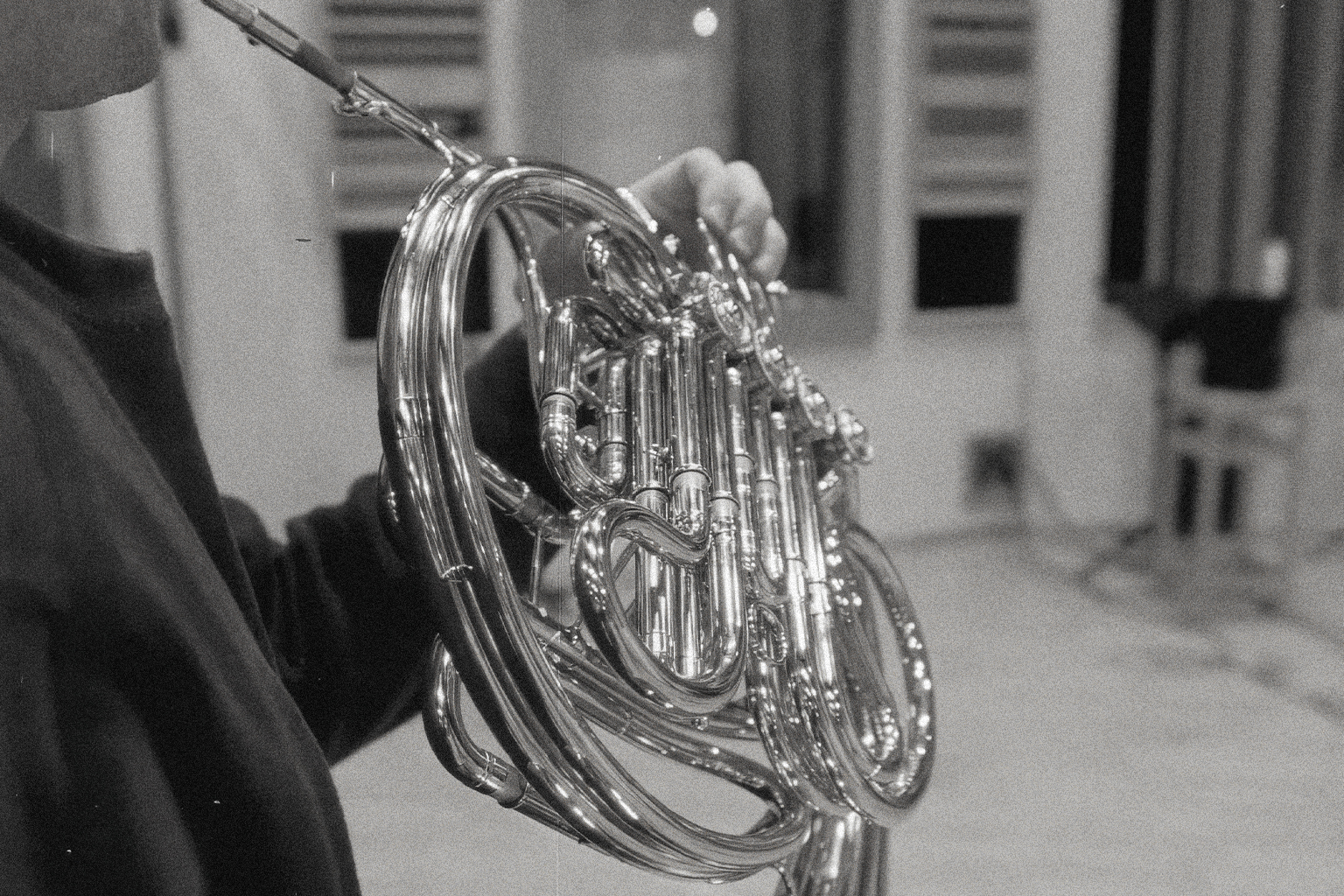 Black and white close-up of a person playing a french horn, showing the instrument's shiny metal tubing and valves.